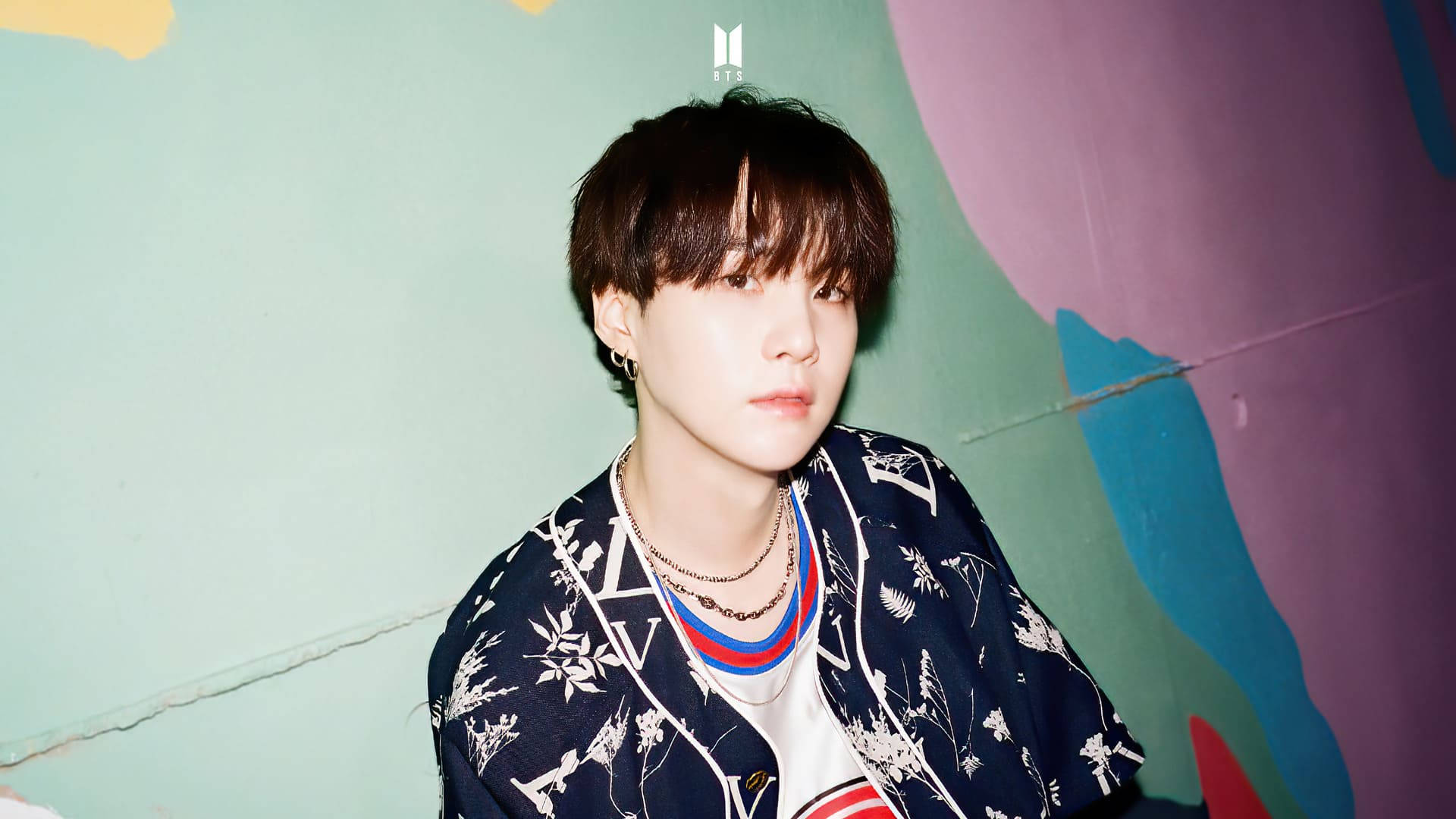 BTS SUGA Dynamite 壁紙 — 一名黑髮、戴著多層項鍊的年輕男子平靜地凝視著一面色彩繽紛的抽象牆壁，帶有青綠色、紫色和藍色調。