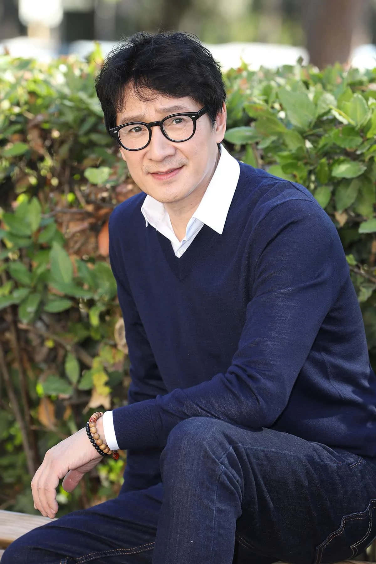 Ke Huy Quan 桌布 壁紙 — 一名黑髮戴眼鏡的男子，靜靜地坐在長椅上，周圍是茂密的綠色灌木叢。