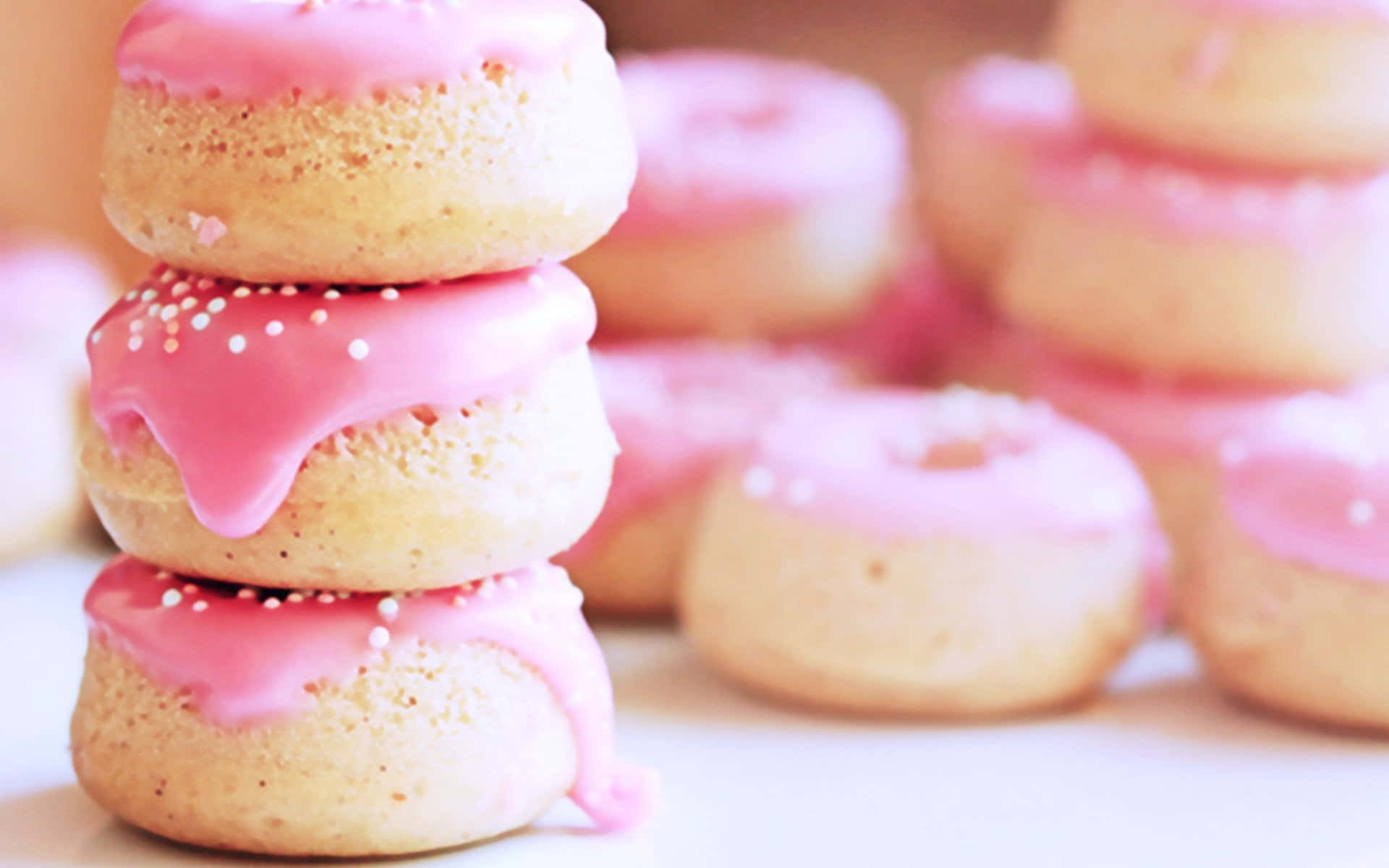 Cute Donut 桌布 壁紙 — 一堆三個粉紅色糖霜甜甜圈，上面有白色糖粒，在柔焦中呈現，背景是模糊的更多甜甜圈。