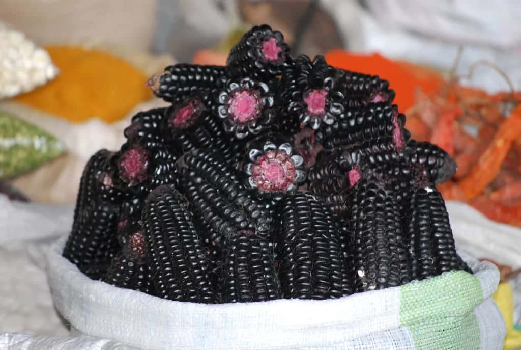Purple Corn 桌布 壁紙 — 一堆帶有粉紅色花心的光澤黑粟米棒放在鋪有白布的籃子裡。