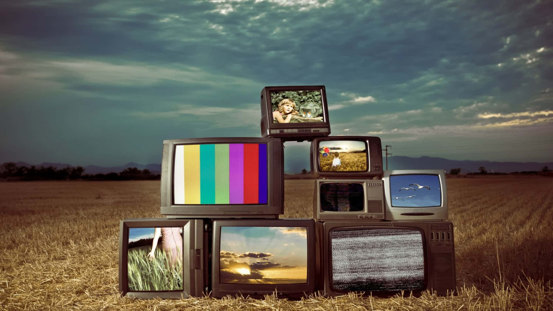 Television Set 桌布 壁紙 — 一堆復古電視機在田野中，在陰沉天空下顯示彩色測試圖案和懷舊場景。