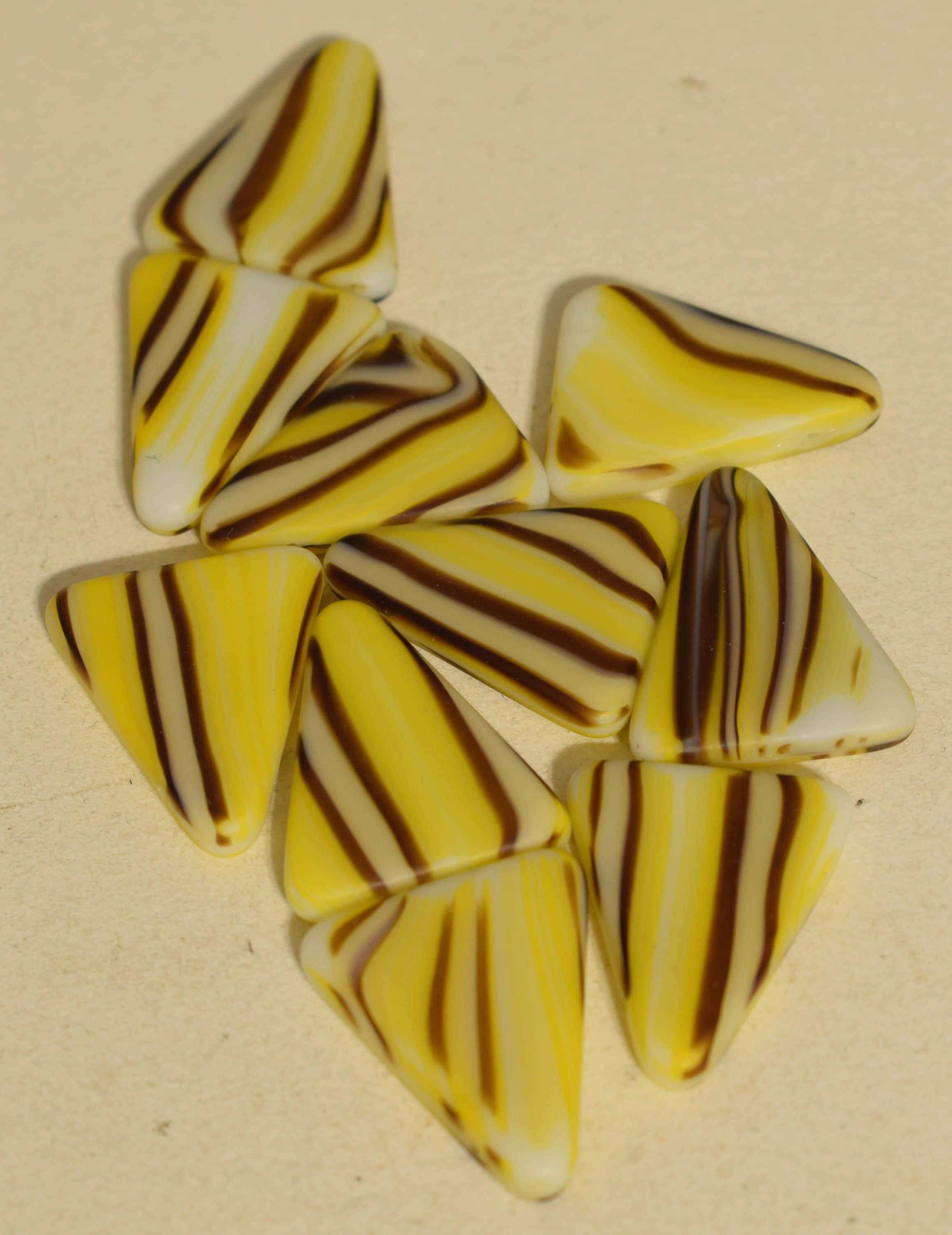 Yellow Triangle 桌布 壁紙 — 一堆散落的光澤三角形玻璃珠，帶有鮮明的黃色、棕色和白色條紋，位於中性米色表面上。
