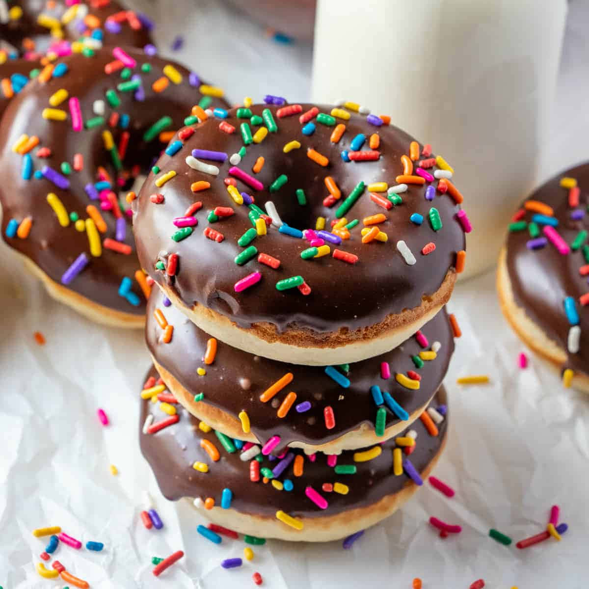 Glazed Donut 桌布 壁紙 — 一堆朱古力釉面冬甩，上面灑滿彩虹糖針，放在皺巴巴的牛油紙上，旁邊有一杯牛奶。