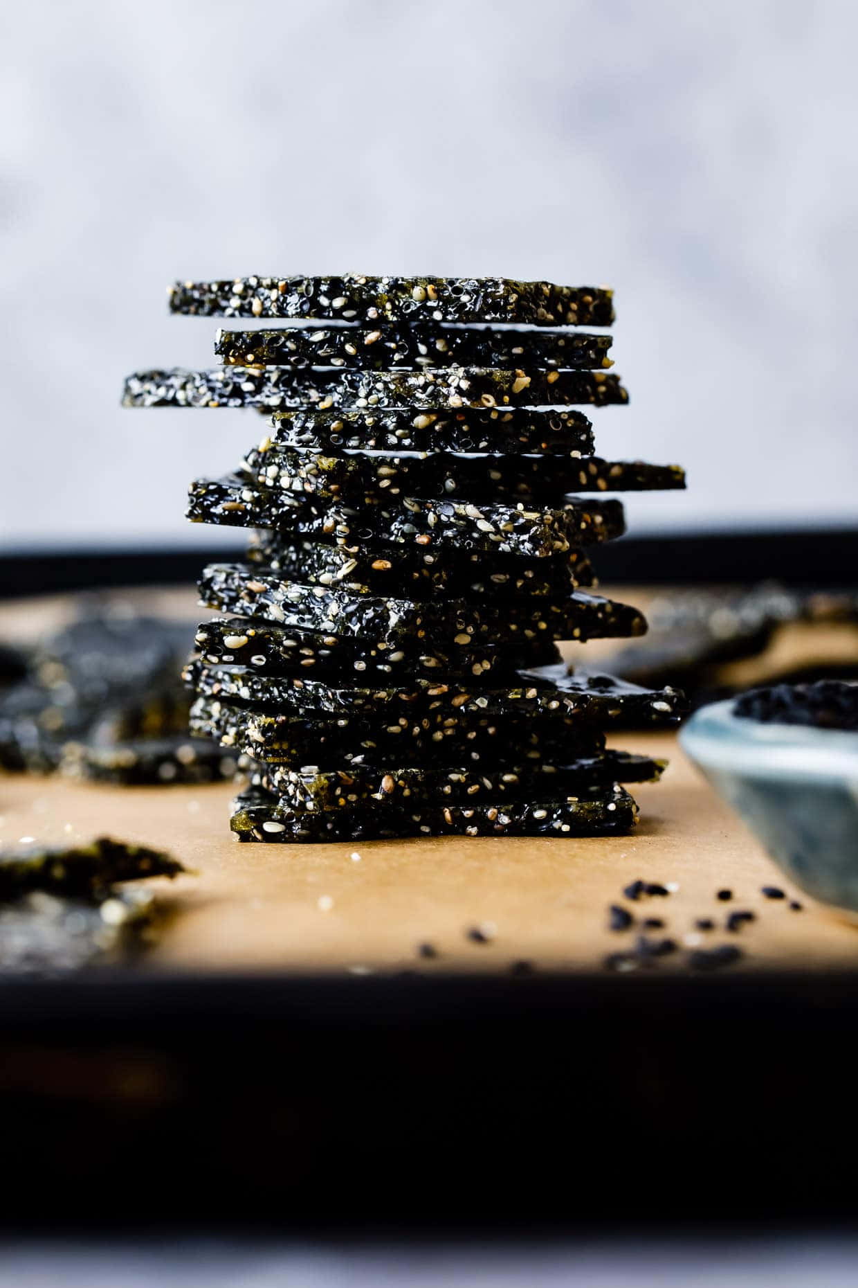 Black Sesame 桌布 壁紙 — 一堆深色芝麻餅乾高高疊起放在木板上，附近有散落的芝麻和一個小碗。