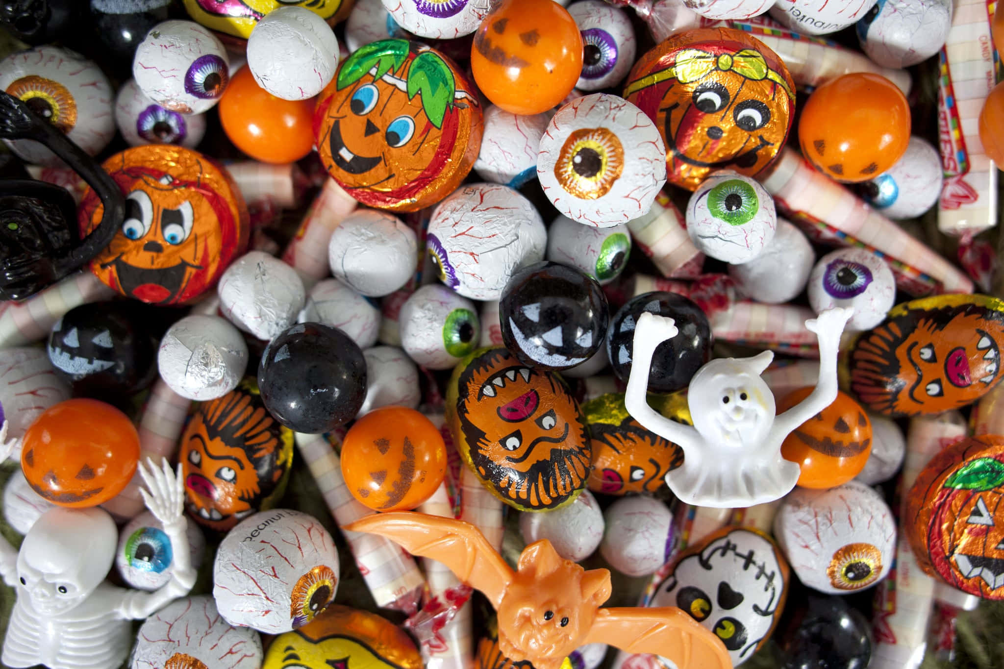 Halloween Treats 桌布 壁紙 — 一堆混亂的萬聖節糖果和玩具，包括眼球、南瓜、鬼魂和蝙蝠，顏色為鮮豔的橙色、白色和黑色。