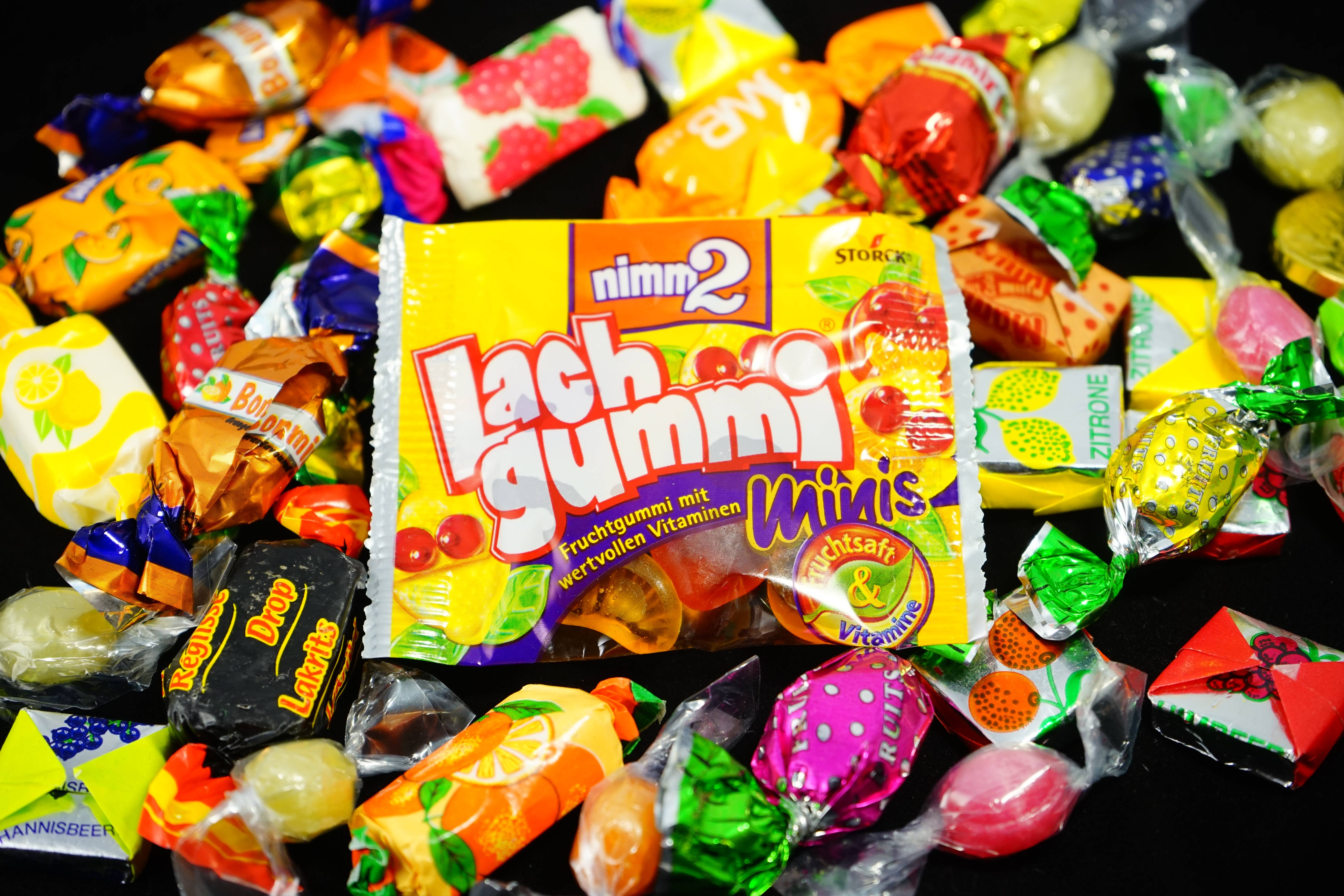 Chewy Lachgummi 壁紙 — 一堆色彩繽紛的包裝糖果圍繞著一個亮黃色的Lachgummi Minis包裝，背景為黑色。