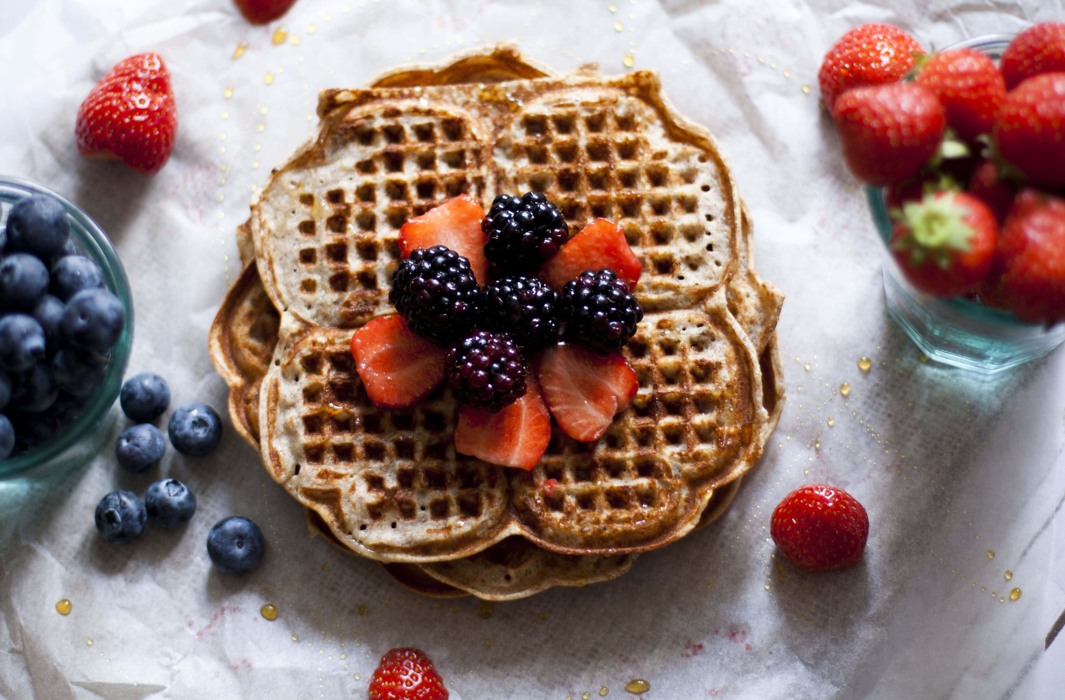 Waffle 桌布 壁紙 — 一堆金黃色窩夫，上面放上新鮮黑莓和草莓，周圍有藍莓和原粒草莓，放在牛油紙上。