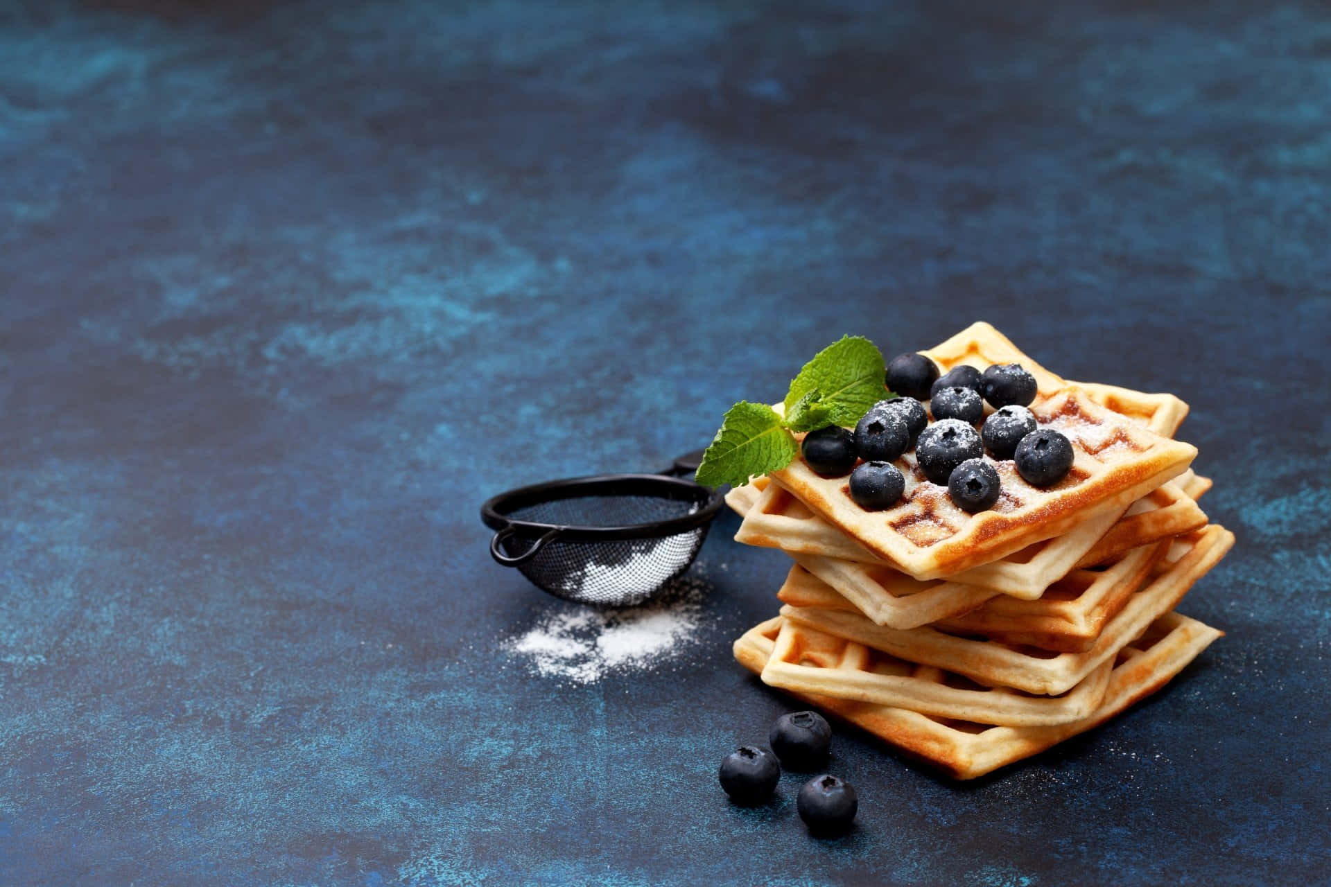 Waffle 桌布 壁紙 — 一堆金黃色窩夫，上面放有藍莓和薄荷，置於有紋理的藍色表面上，並撒有糖粉。