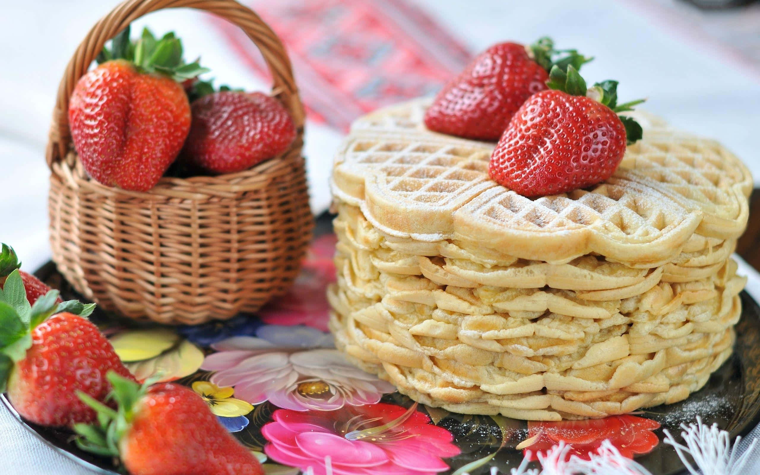 Waffle 桌布 壁紙 — 一堆高高的金黃色窩夫，撒上糖霜，上面放上新鮮草莓，旁邊有一個編織籃子，裡面有更多草莓，放在一個花卉圖案的碟子上。