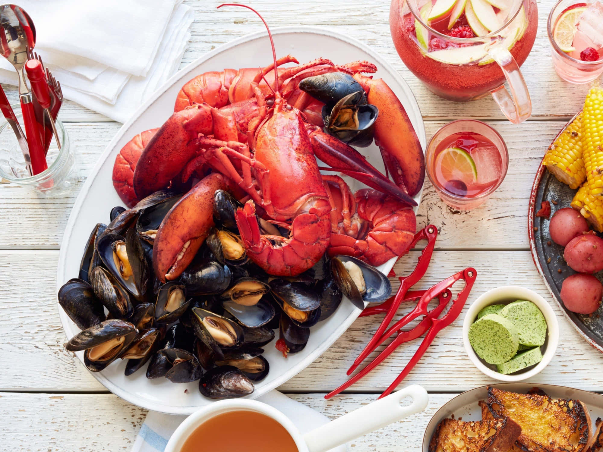 Seafood 桌布 壁紙 — 一場充滿活力的海鮮盛宴，有鮮紅色的龍蝦和黑色青口放在白色碟上，周圍有粟米、薯仔和清涼飲品，放在鄉村風格的木桌上。