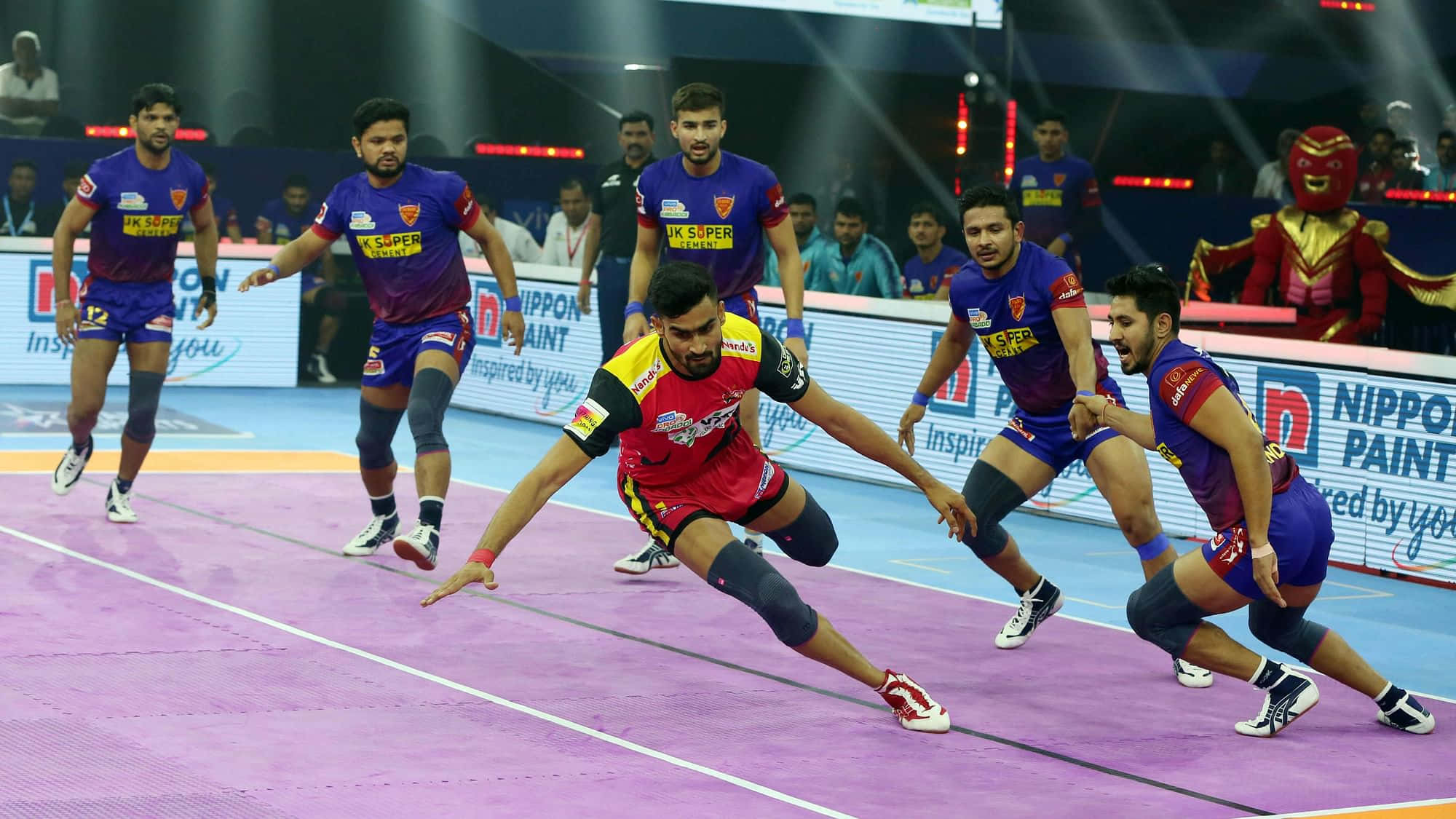 Kabaddi 桌布 壁紙 — 一場動態的板網球比賽展開，紅隊球員低身撲球，而紫隊防守球員在明亮的場館燈光下逼近。