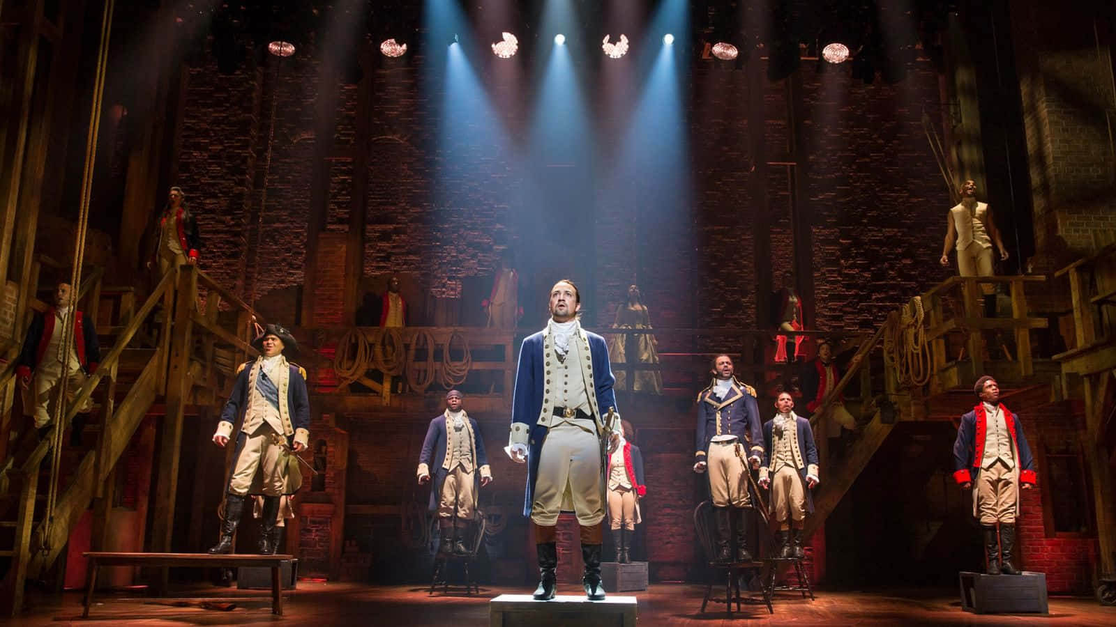 Hamilton 桌布 壁紙 — 一場戲劇性的舞台表演，演員穿著殖民時期制服，在明亮的聚光燈下，於質樸的木製佈景中演出。