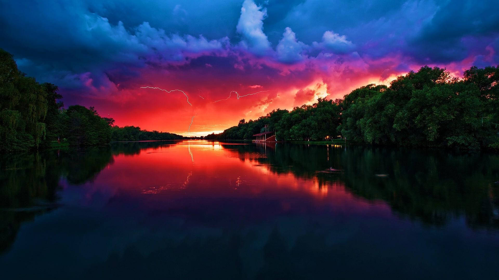 自然 桌布 壁紙 — 一場戲劇性的風暴，伴隨著鮮豔的紅色和紫色閃電，照亮了一條被茂密綠樹環繞的平靜河流。