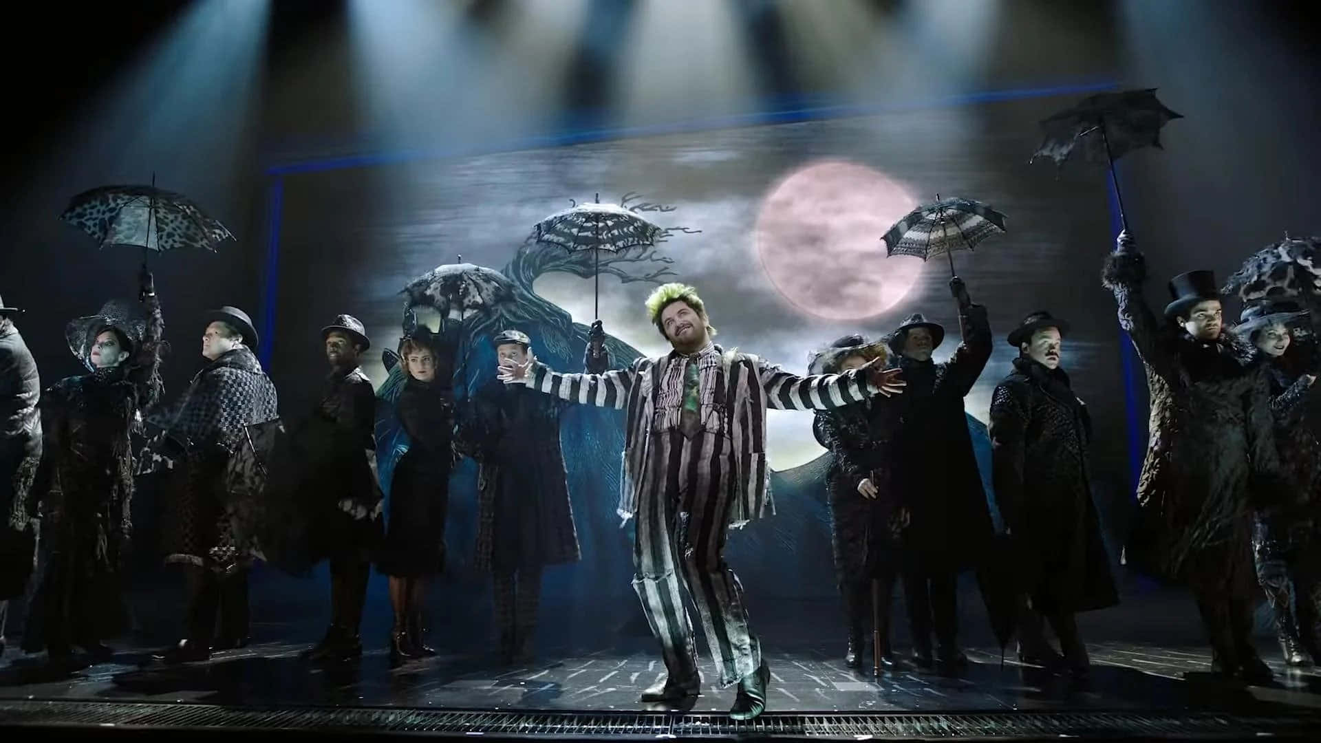 Beetlejuice 桌布 壁紙 — 一場戲劇舞台表演，中央有一名綠色頭髮的條紋人物，周圍是穿著戲服的演員，在戲劇性聚光燈下撐著雨傘。