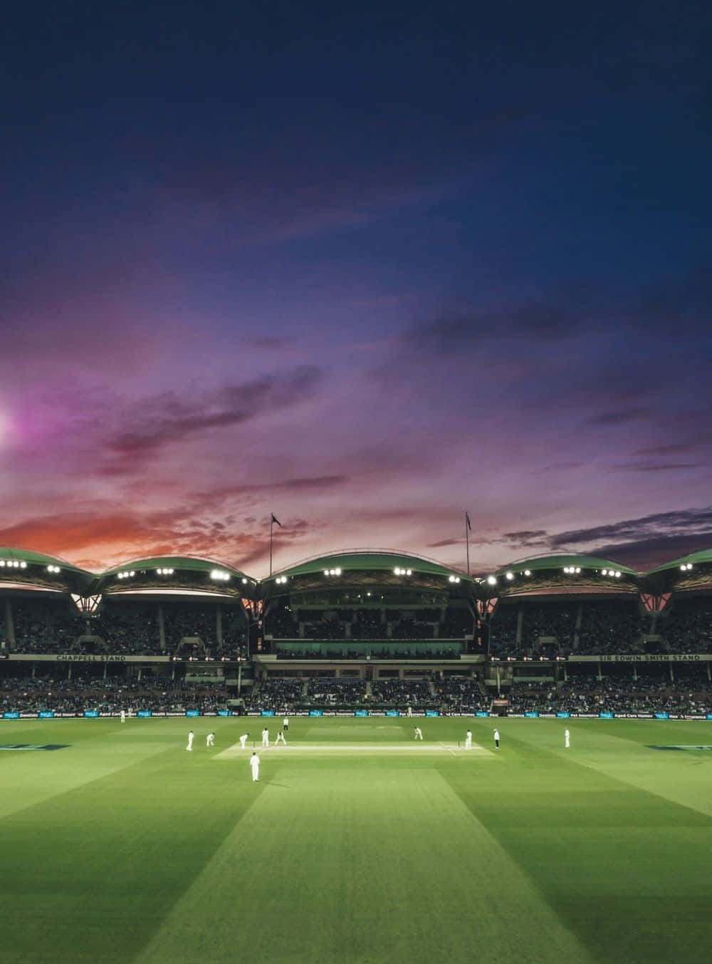 Cricket 桌布 壁紙 — 一場板球比賽在充滿活力的暮色天空下展開，體育場的燈光照亮了綠色的場地和座無虛席的看台。