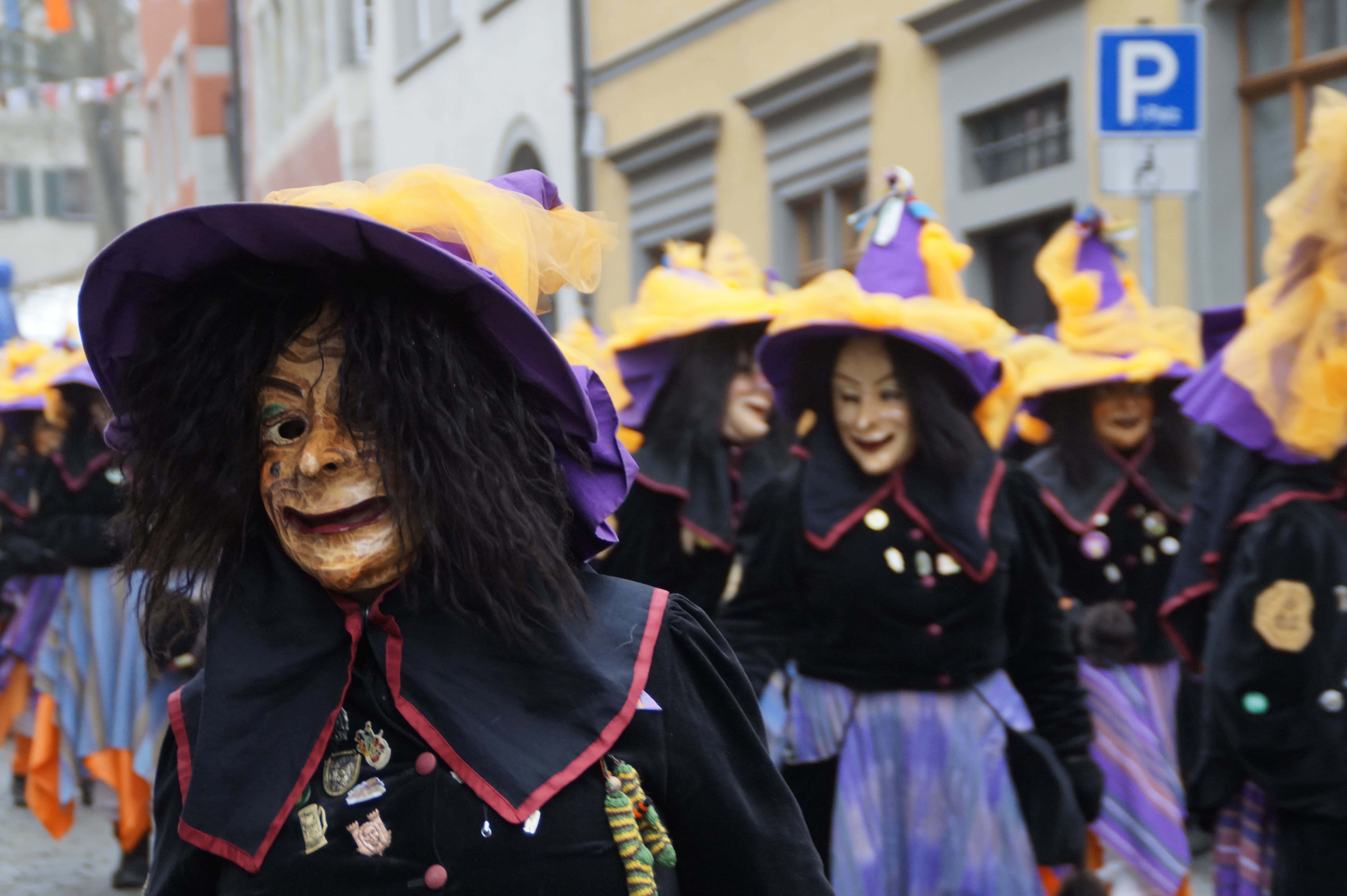 Halloween Parades 桌布 壁紙 — 一場熱鬧的街頭遊行，打扮成女巫的人們戴著紫色帽子、臉上塗著顏料，在歐洲小鎮中遊行。