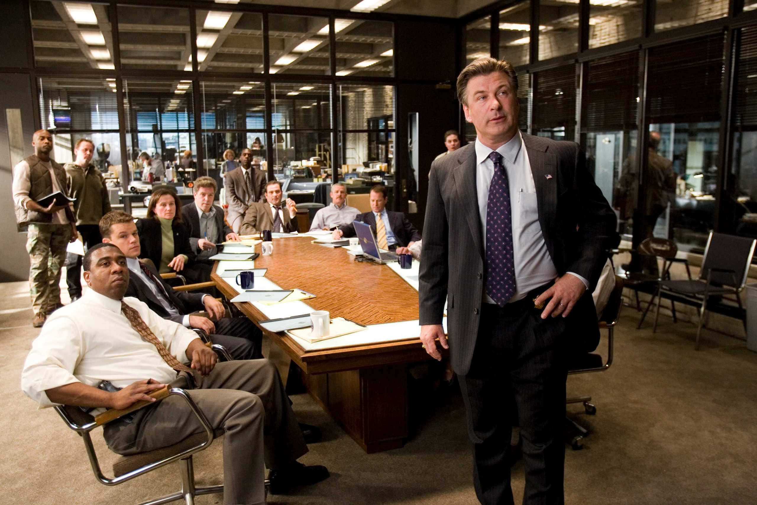 Alec Baldwin 桌布 壁紙 — 一場緊張的企業會議在現代玻璃牆辦公室中展開，身穿西裝的男士們圍坐在一張長木桌旁。