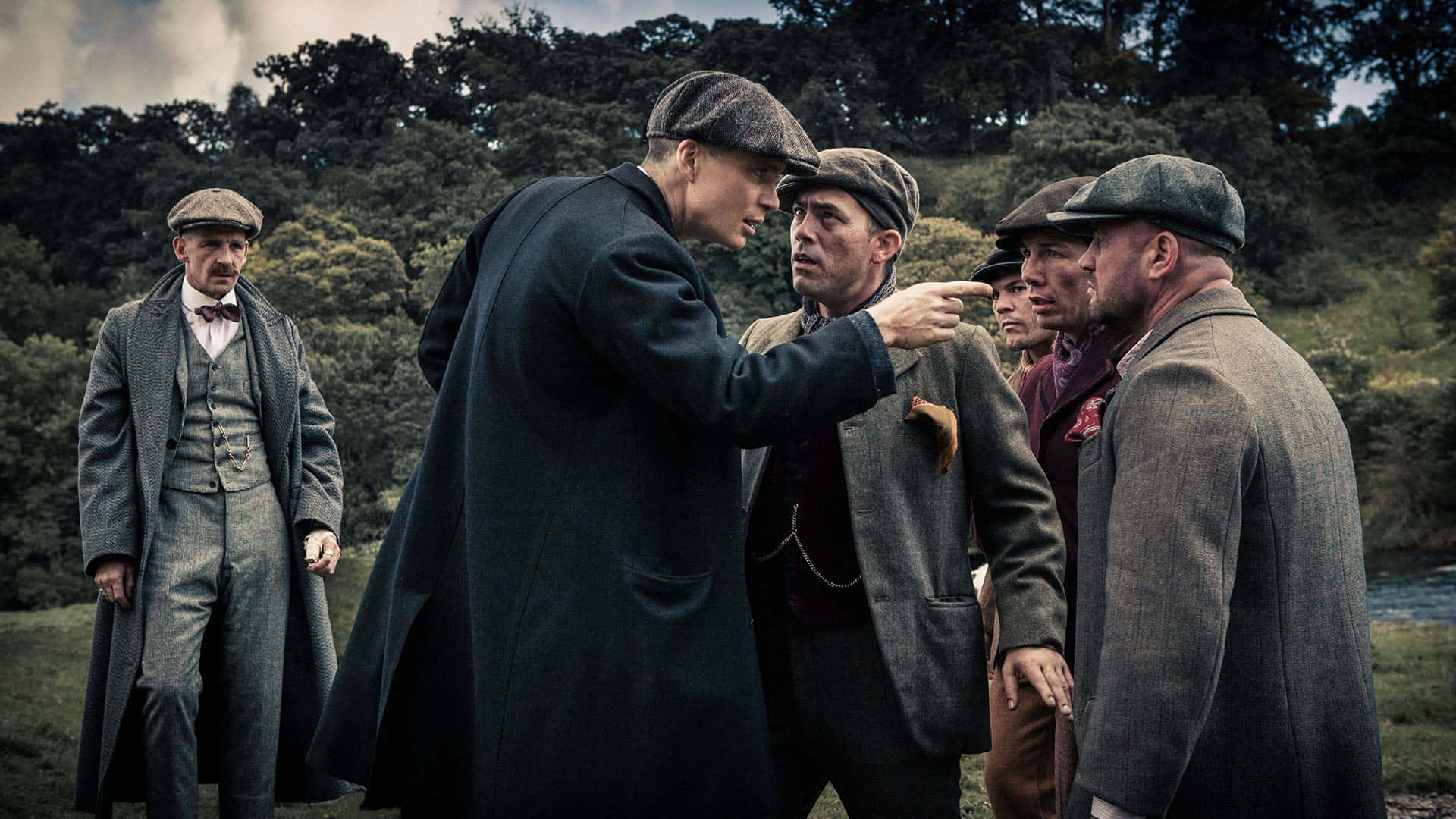 Peaky Blinders 桌布 壁紙 — 一場緊張的對峙展開，一名身穿深色外套的男子指責性地指向一群五名穿著復古粗花呢西裝和扁平帽子的男子中的另一名男子。