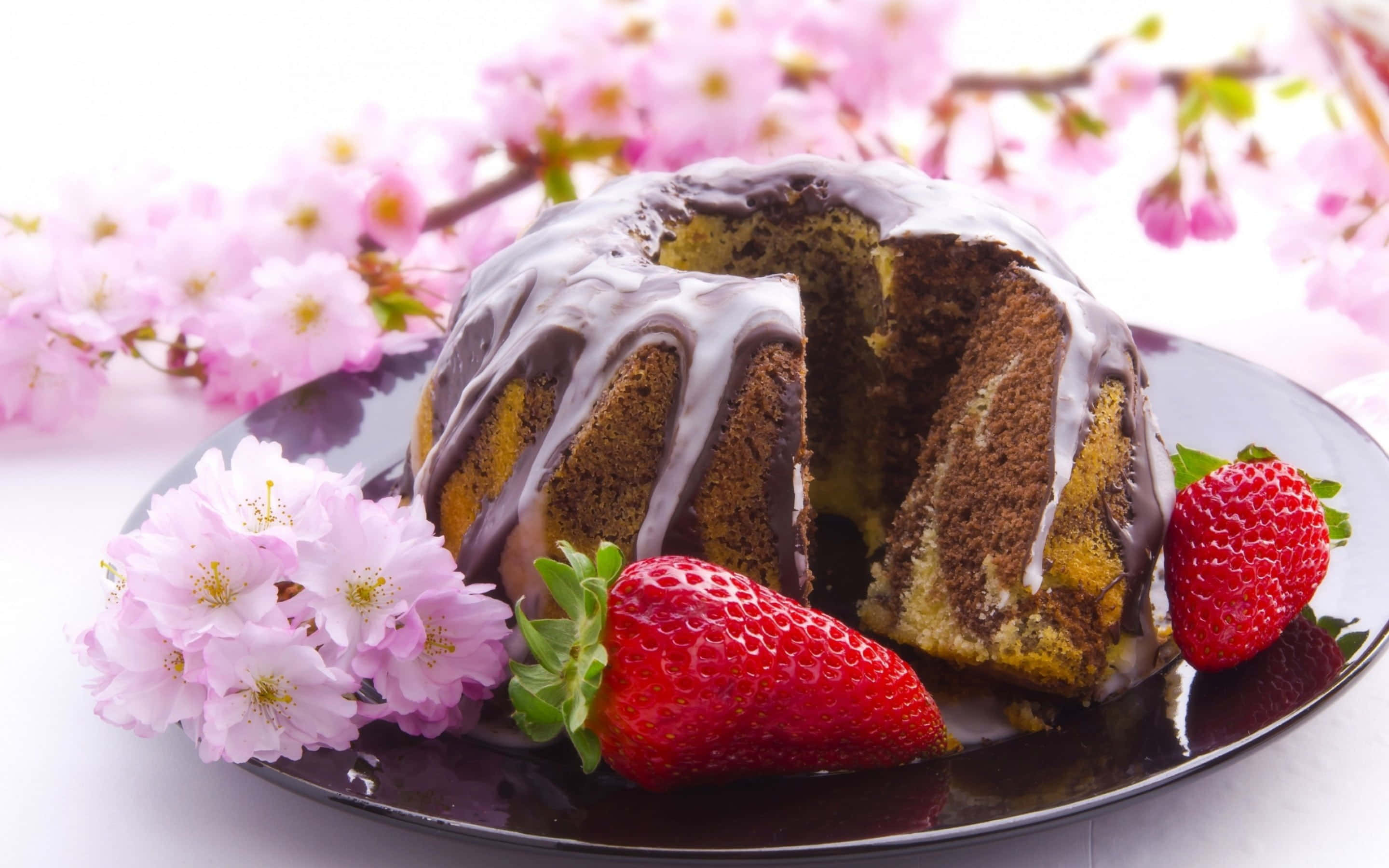 Spring Desserts 桌布 壁紙 — 一塊切片朱古力環形蛋糕配白色糖霜，放在深色碟上，旁邊有新鮮草莓和粉紅櫻花。