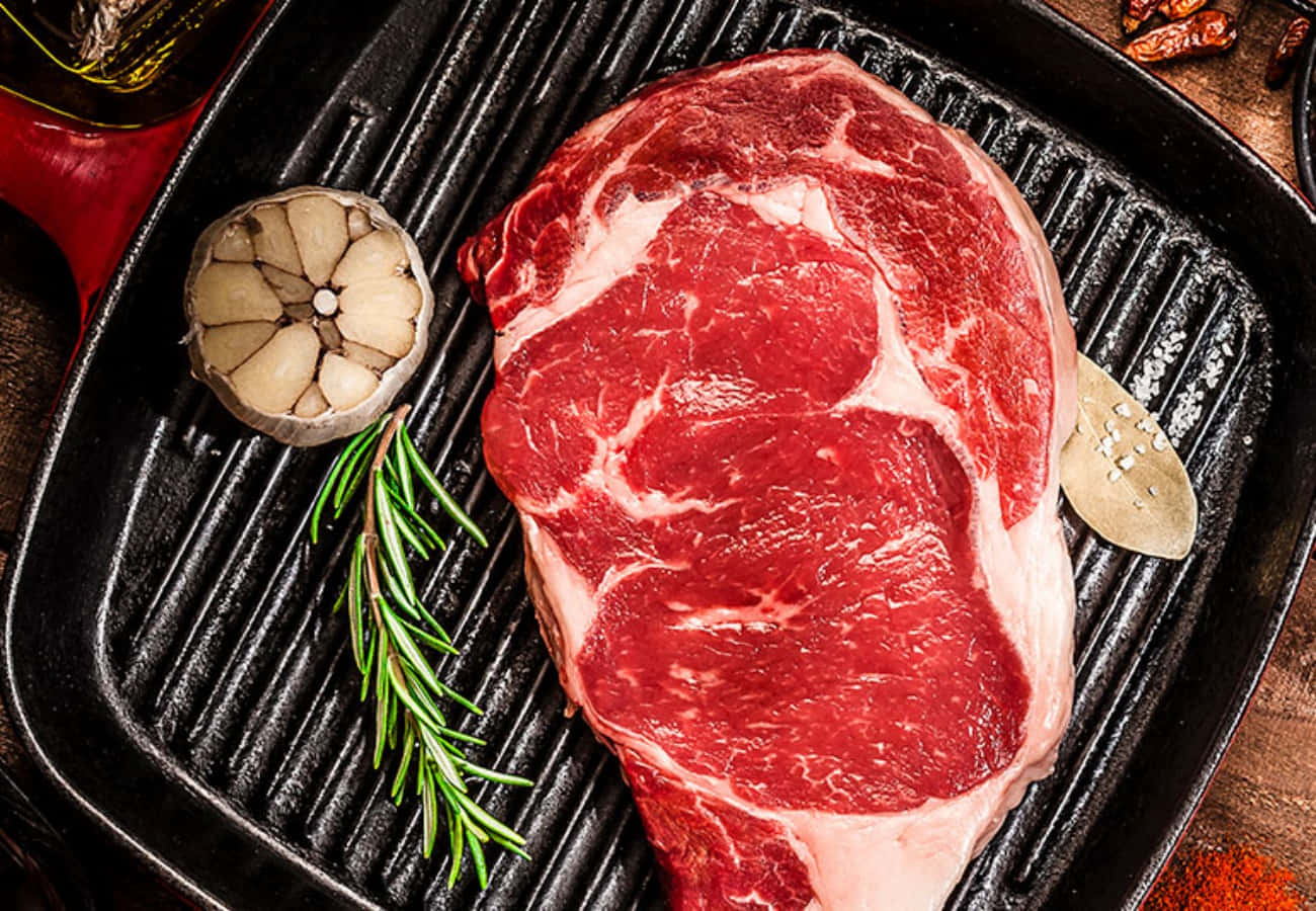 meat 桌布 壁紙 — 一塊多汁的生牛扒放在黑色烤盤上，旁邊有大蒜、迷迭香和月桂葉，準備烹調。