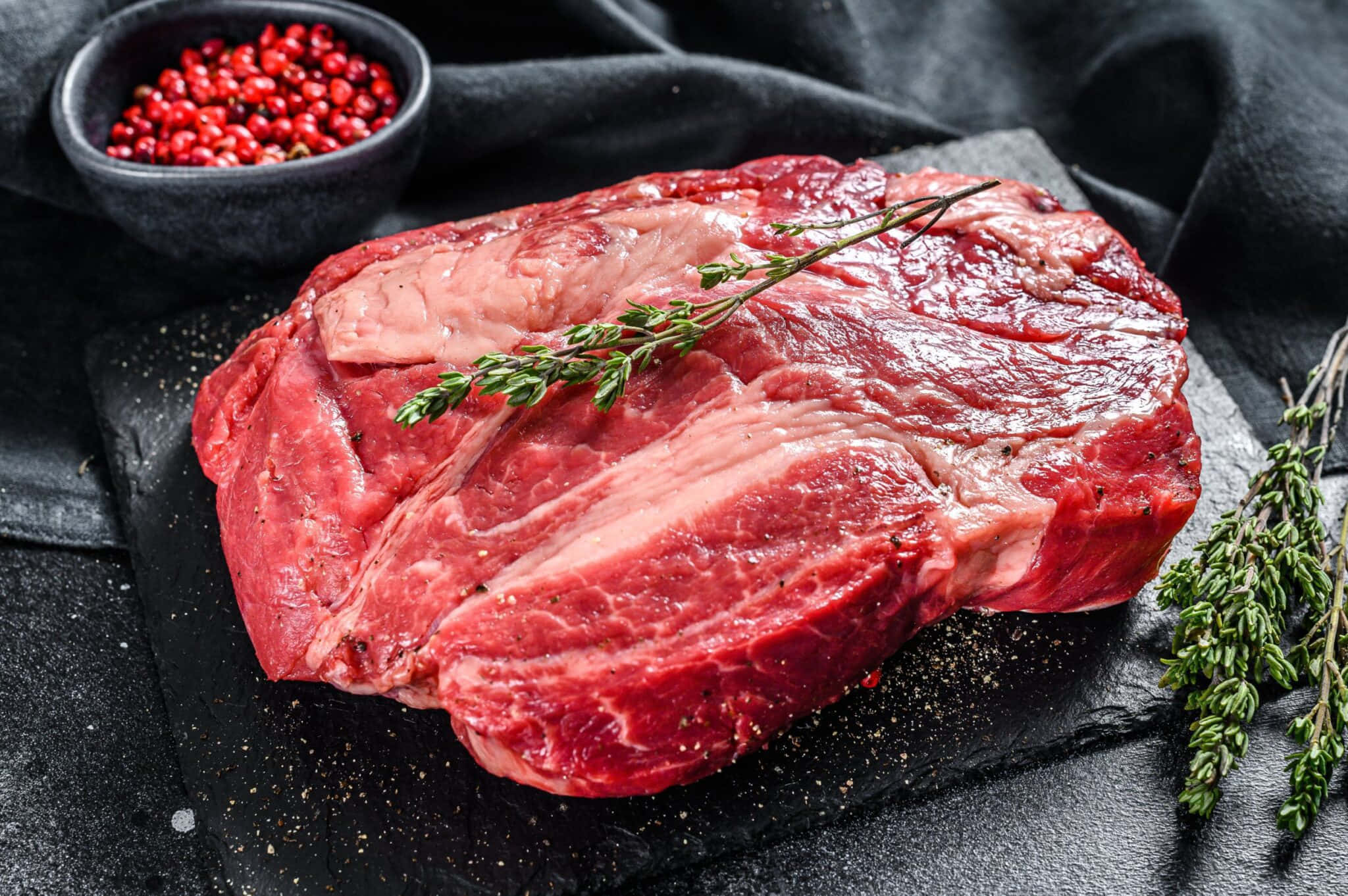 Red Meat 桌布 壁紙 — 一塊多汁的生牛肉扒放在深色石板板上，飾以新鮮百里香和紅胡椒粒。