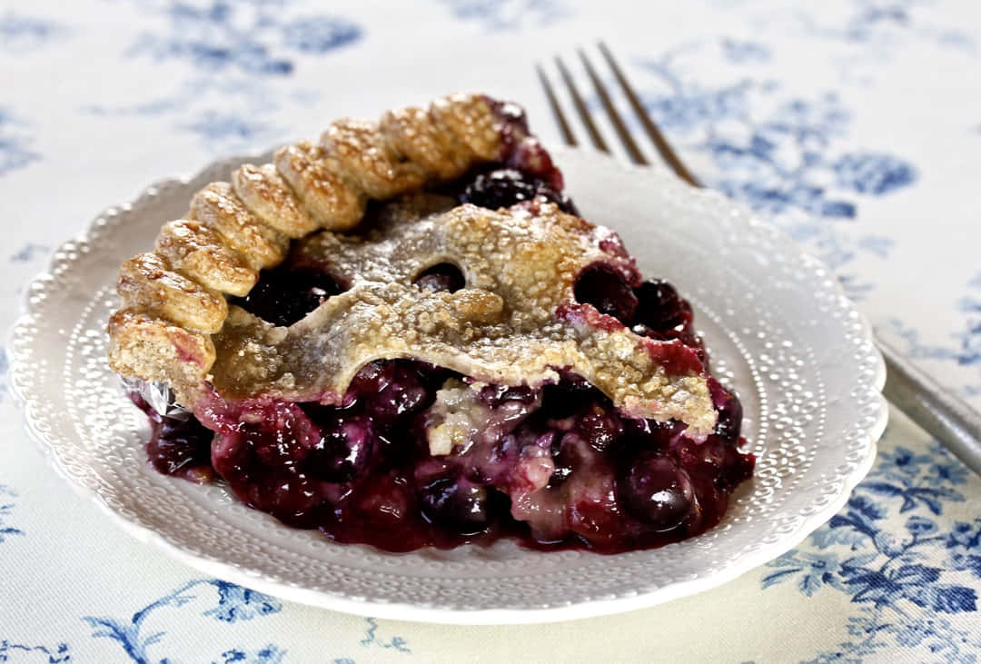 Blueberry Pie 桌布 壁紙 — 一塊多汁的藍莓批，帶有金黃酥脆的外皮，放在白色花紋碟上，旁邊有一把叉。