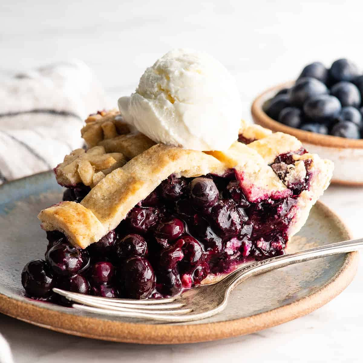 Blueberry Pie 桌布 壁紙 — 一塊多汁的藍莓批，配上酥脆的外皮和雲呢拿雪糕，放在一個鄉村風格的碟子上，旁邊有叉子和一碗新鮮莓果。