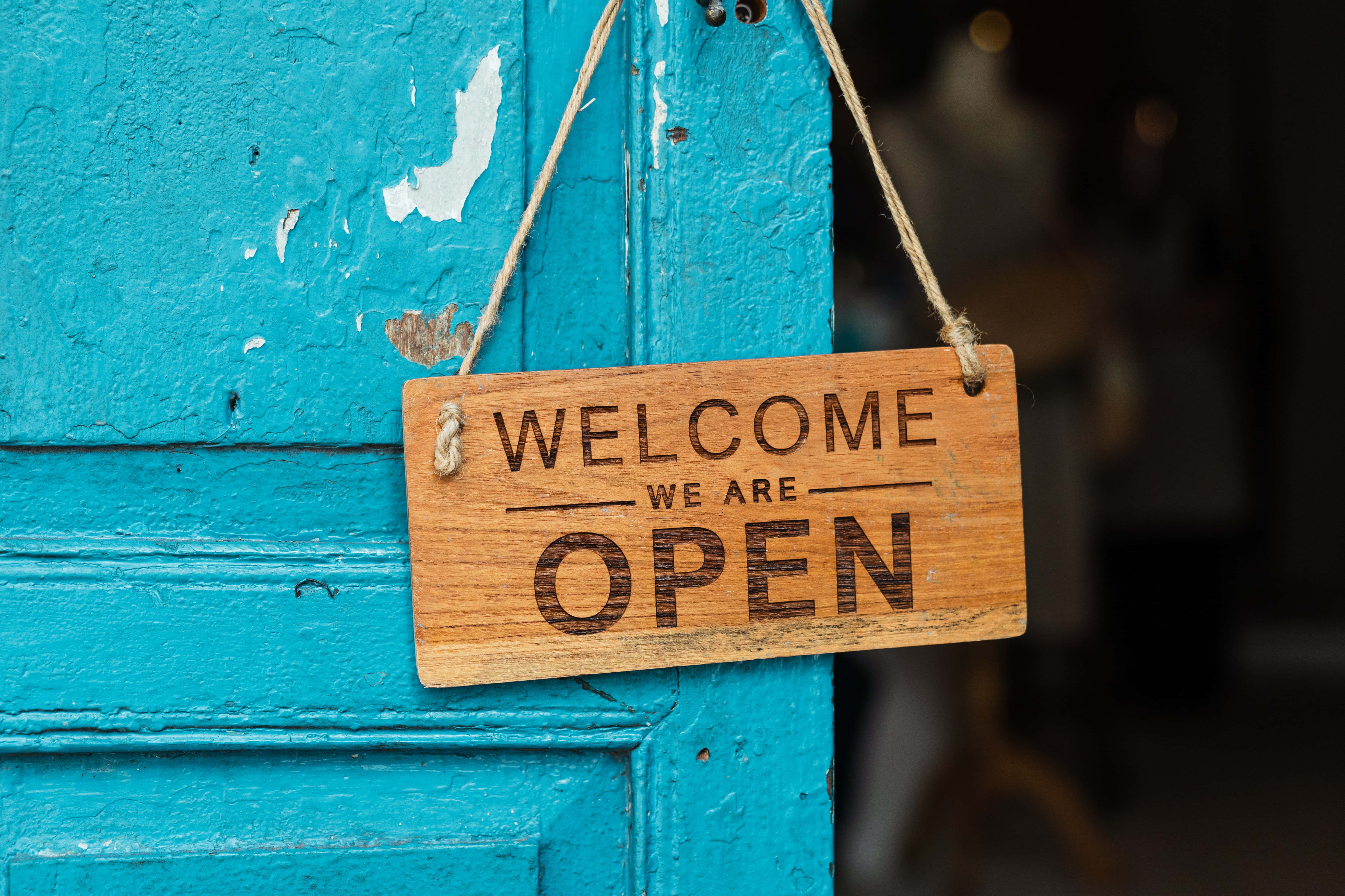 Welcome 桌布 壁紙 — 一塊寫有「WELCOME WE ARE OPEN」的質樸木牌用麻繩掛在褪色剝落的青綠色門上。