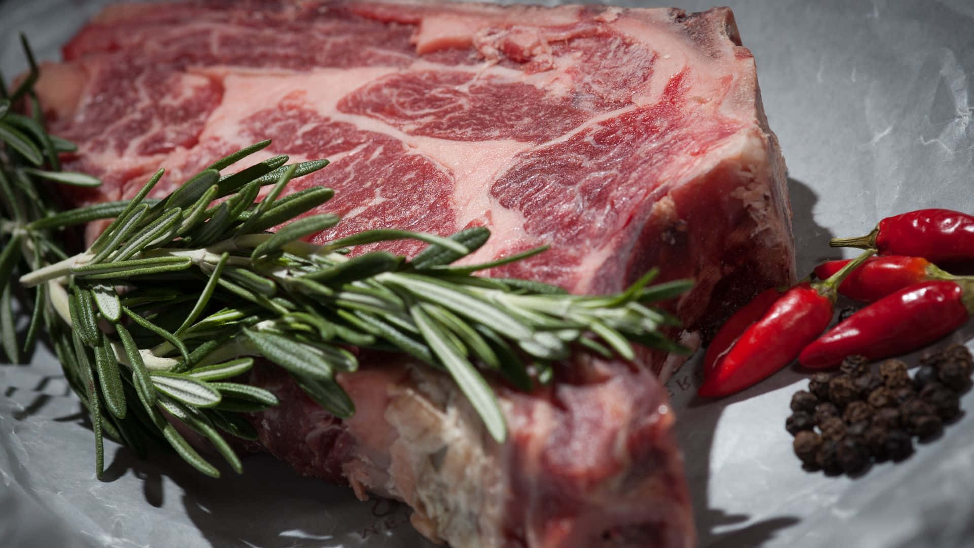 Red Meat 桌布 壁紙 — 一塊帶有大理石花紋的生牛肉扒放在錫紙上，旁邊有迷迭香、紅辣椒和胡椒粒。