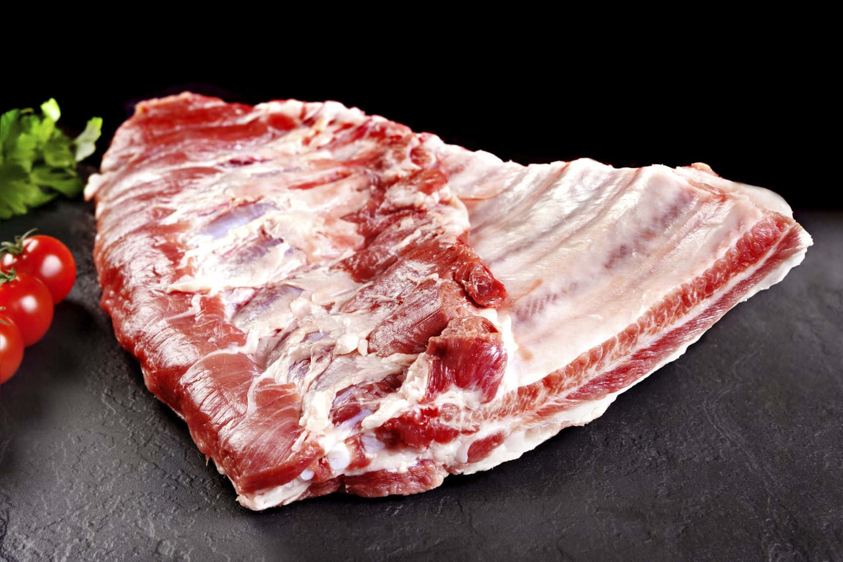 meat 桌布 壁紙 — 一塊帶有大理石花紋的生牛肉肋眼牛排放在深色石板表面上，旁邊是新鮮的車厘茄和歐芹。