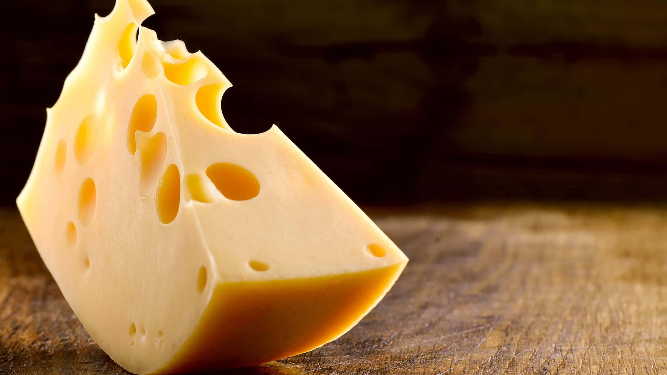 Cheese 桌布 壁紙 — 一塊帶有特徵孔洞的瑞士芝士楔形塊，在溫暖燈光下放置於質樸的木質表面上。