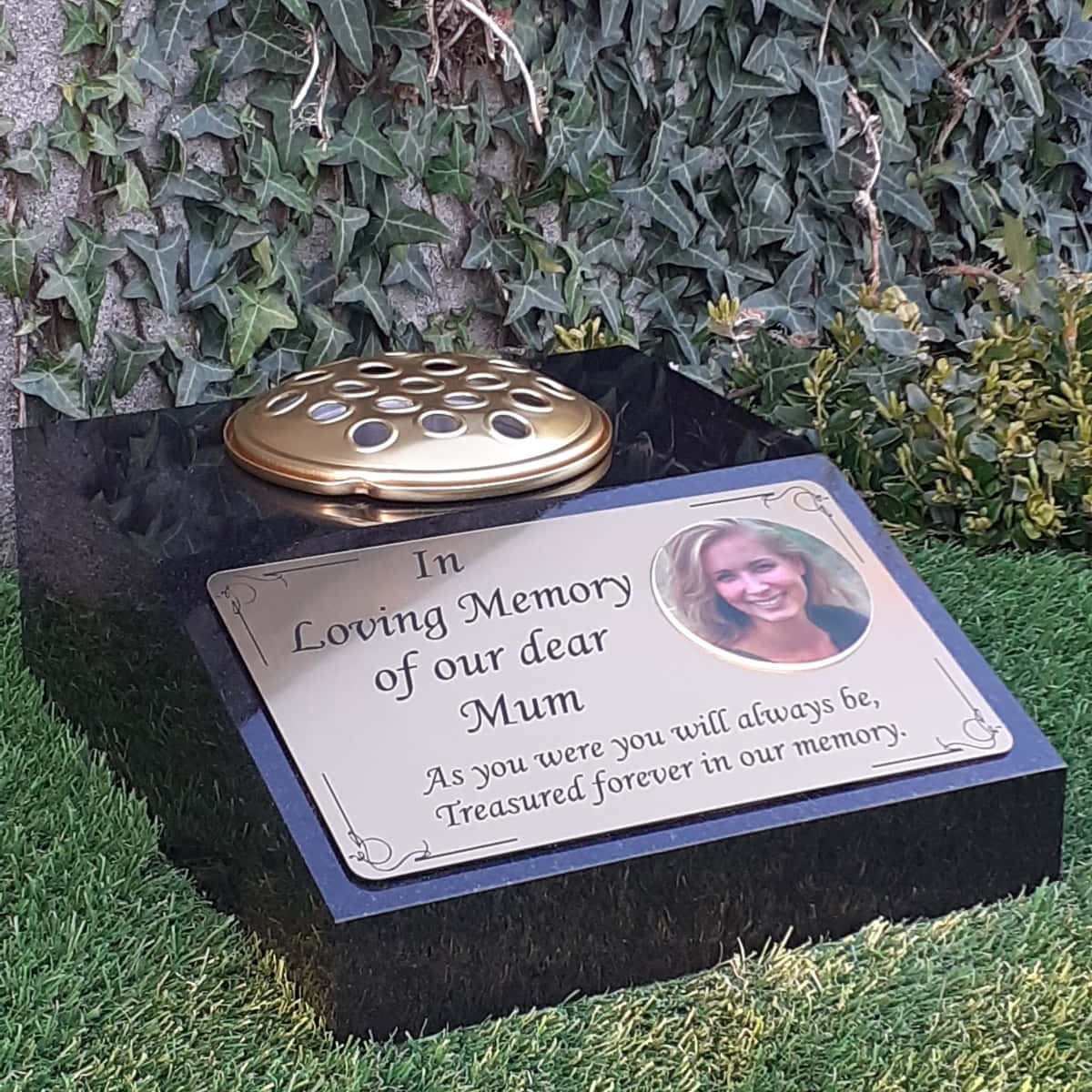 Memorial 桌布 壁紙 — 一塊拋光黑色紀念牌，上面有金色骨灰甕和一位微笑女士的照片，放在草地上，旁邊是常春藤覆蓋的石頭。