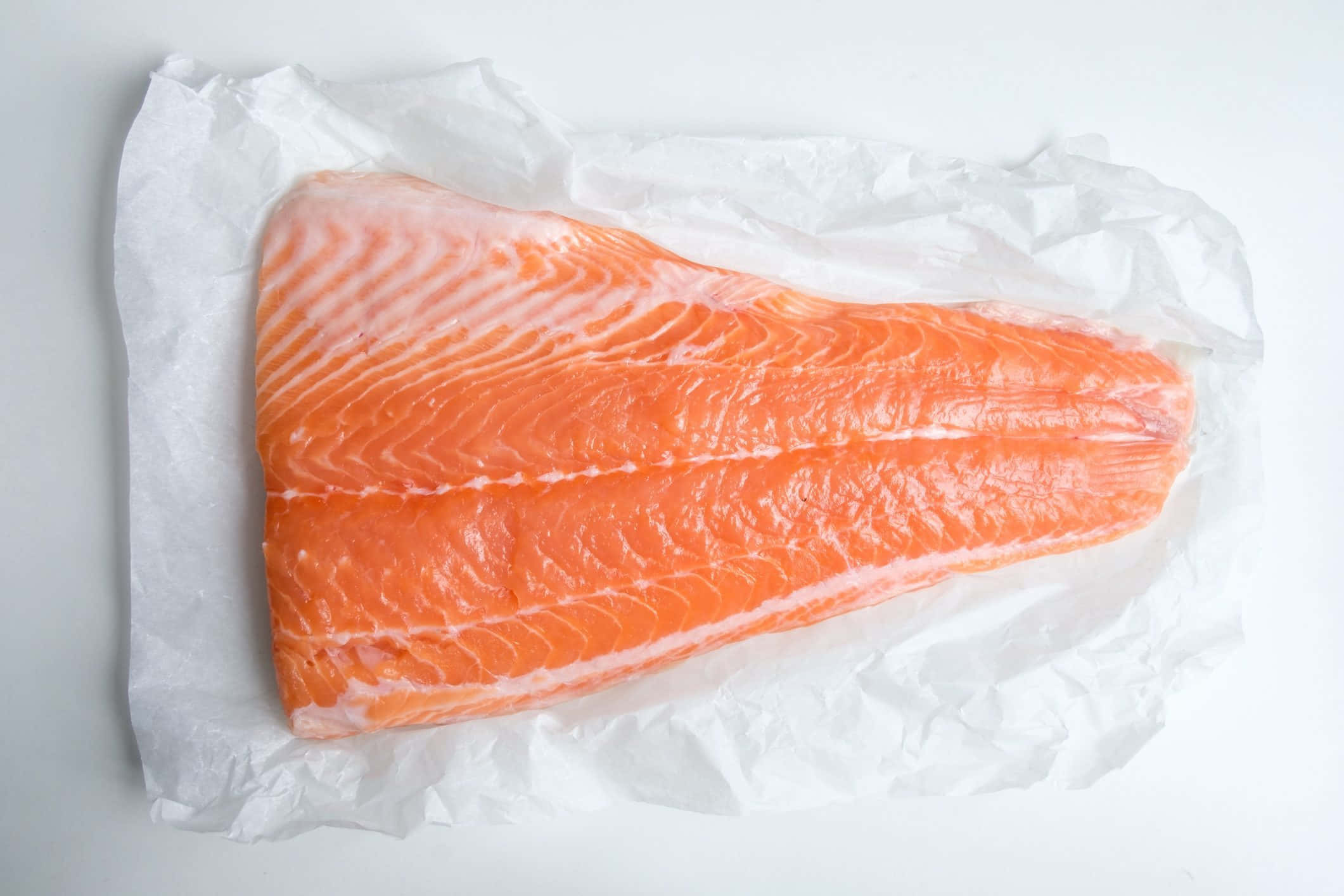 Salmon 桌布 壁紙 — 一塊新鮮的三文魚柳，帶有鮮豔的橙色肉質和白色脂肪紋理，放在皺巴巴的白色牛油紙上，背景是純白色。