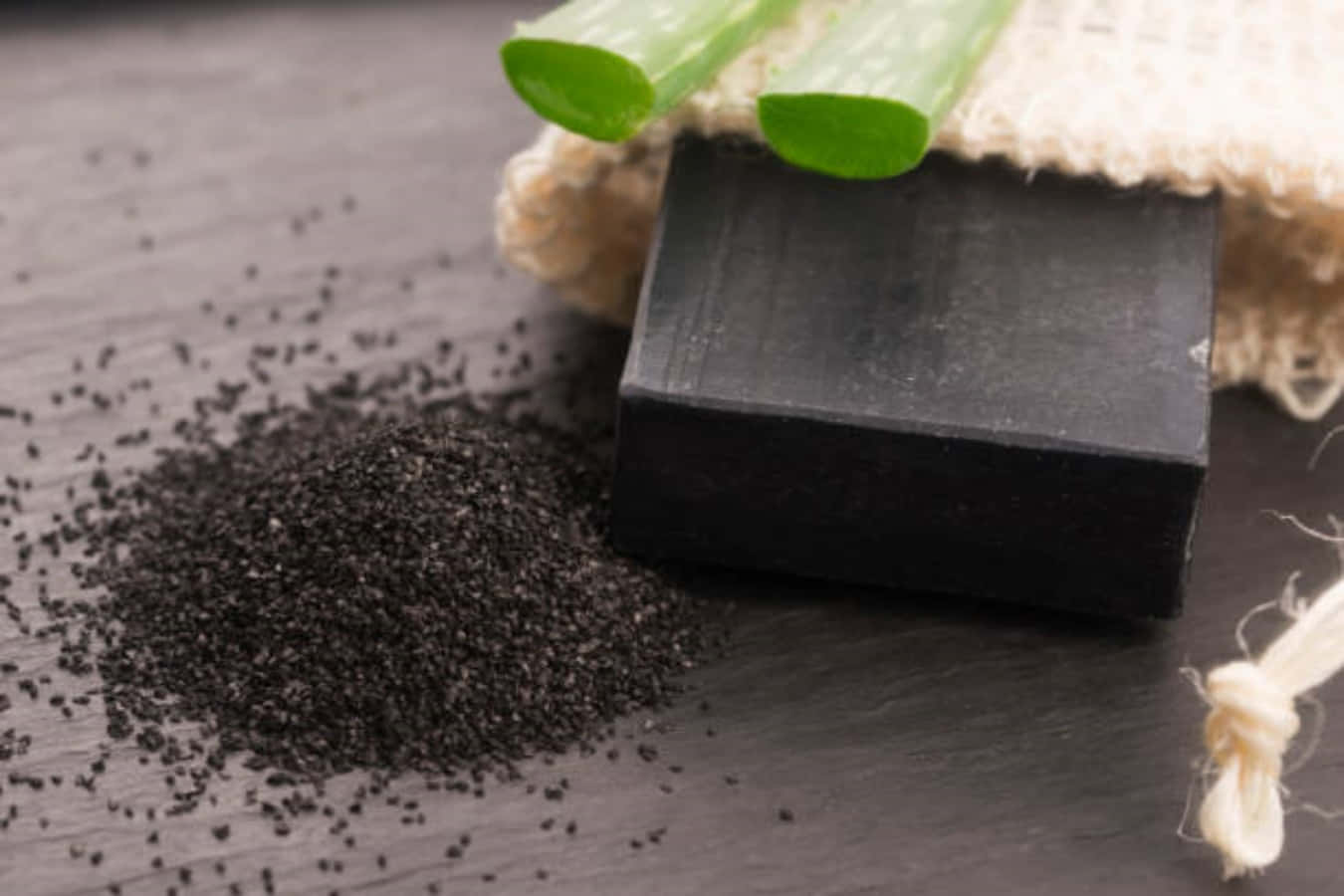 Black Soap 桌布 壁紙 — 一塊時尚的黑色肥皂放在有紋理的石頭表面上，旁邊是一堆深色顆粒和新鮮的青蔥片。