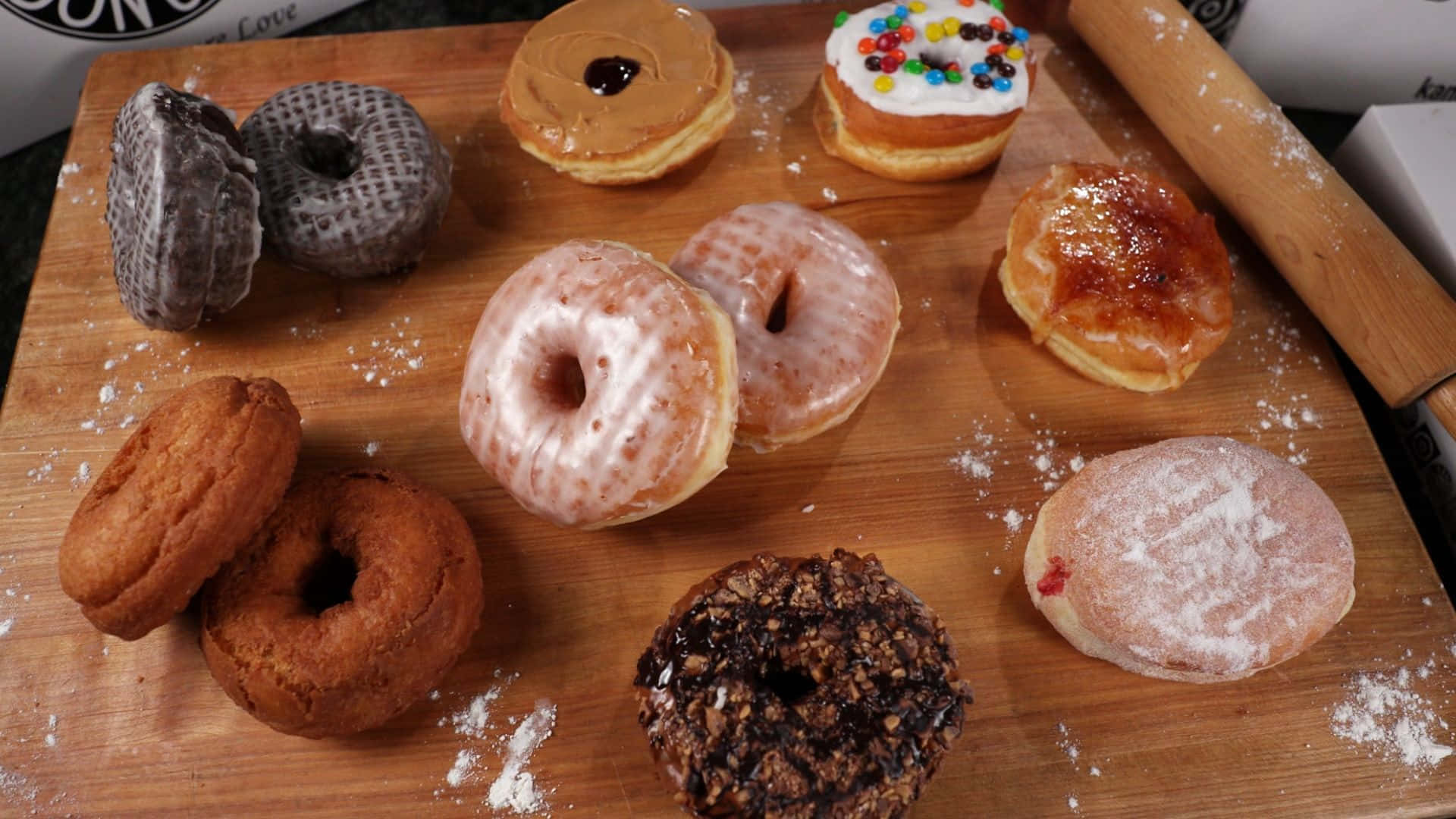 donuts 桌布 壁紙 — 一塊木板上展示著各種帶有糖霜、彩針和糖粉的冬甩，旁邊還有一個擀麵棍。