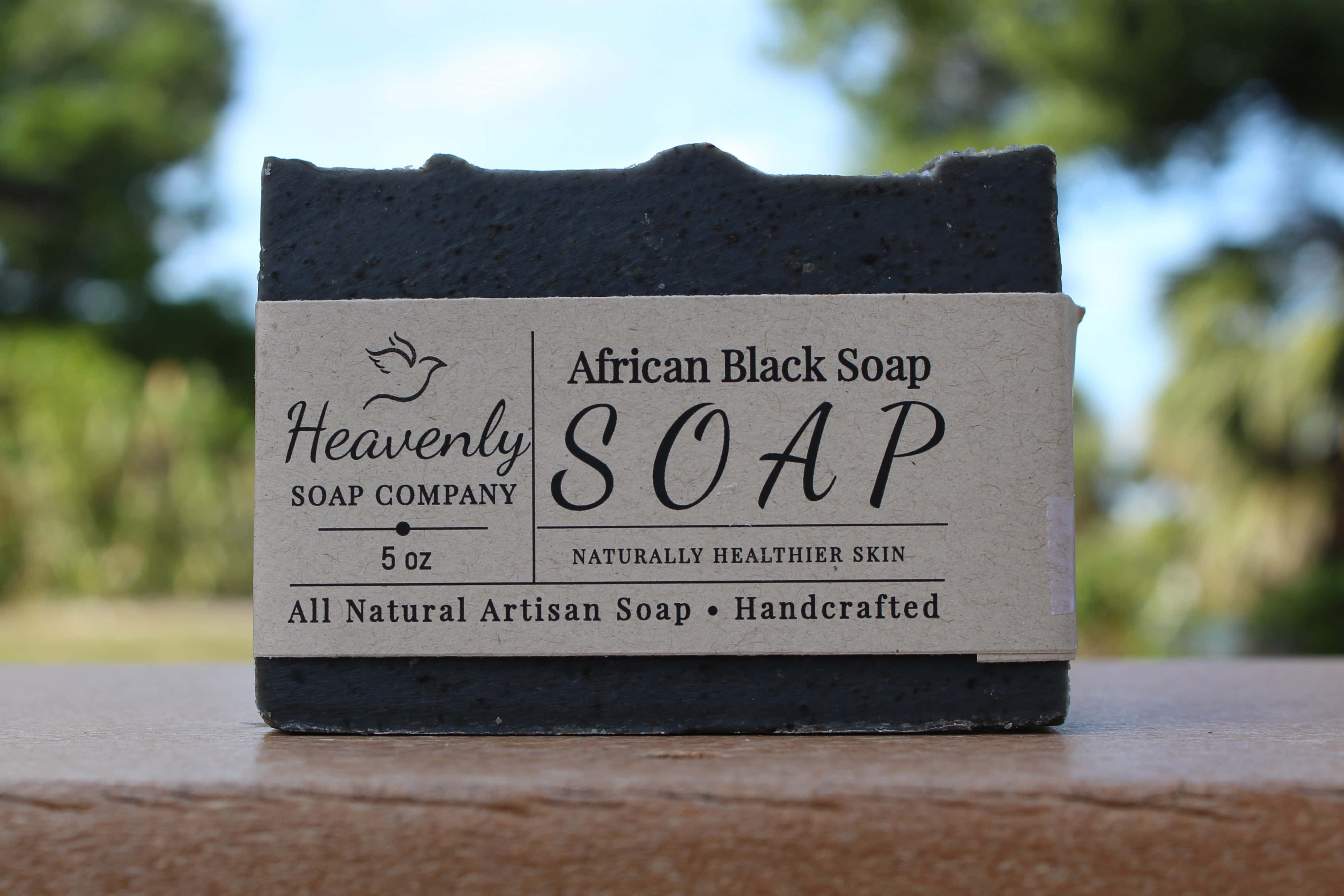 Black Soap 桌布 壁紙 — 一塊深色手工非洲黑肥皂，帶有質樸標籤，放在木質表面上，背景是明亮的綠色戶外。