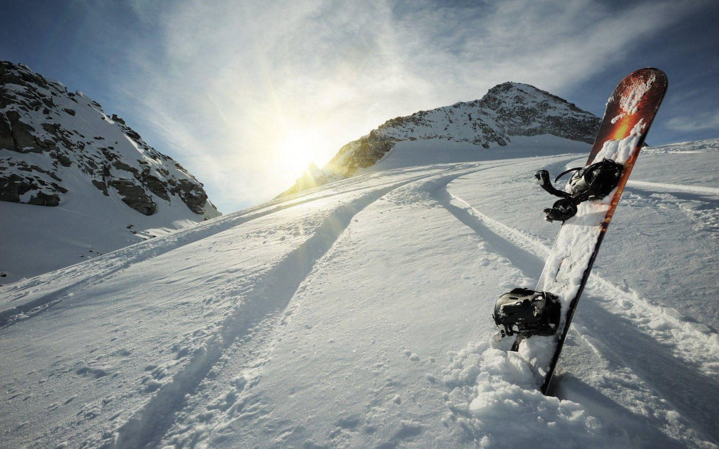 雪 桌布 壁紙 — 一塊滑雪板豎立在新雪上，位於陽光普照的山坡，背景是崎嶇的山峰和明亮的天空。