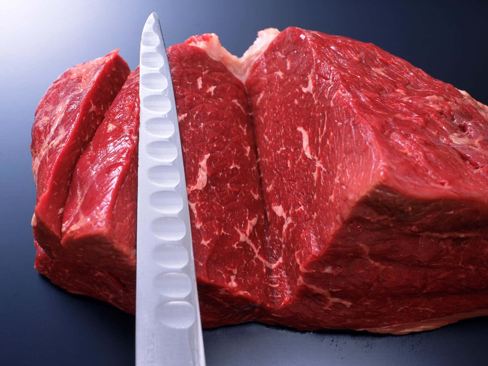 Red Meat 桌布 壁紙 — 一塊生牛肉切塊的特寫，一把鋸齒刀放在深色表面上。
