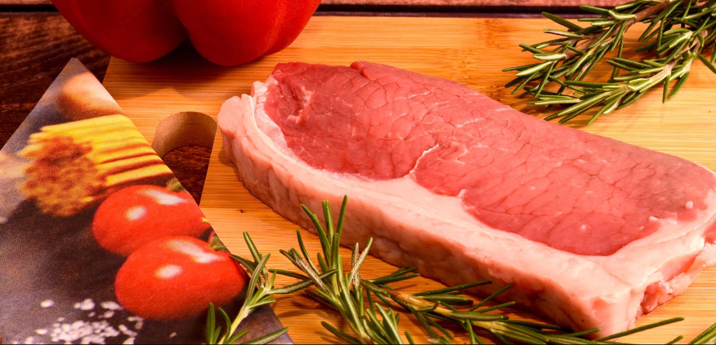 Red Meat 桌布 壁紙 — 一塊生牛肉扒放在木板上，周圍有新鮮迷迭香、車厘茄和紅甜椒。