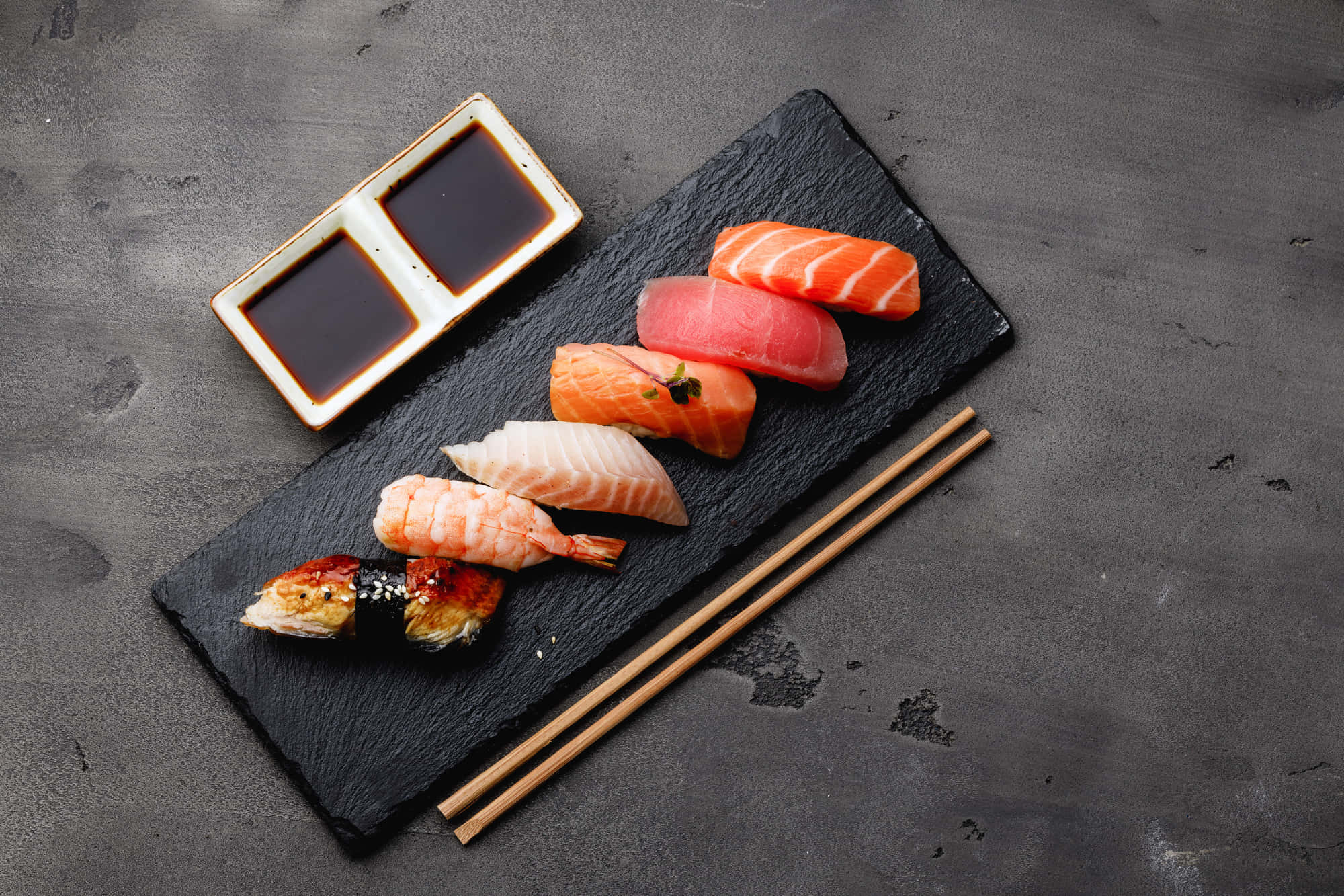 Sushi 桌布 壁紙 — 一塊石板托盤上放著各式握壽司，配以醬油和筷子，置於有紋理的灰色表面上。