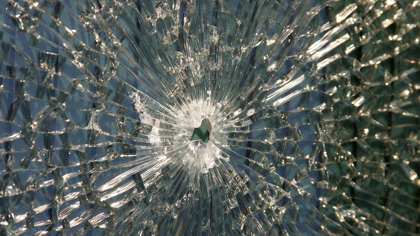 cracked 桌布 壁紙 — 一塊破碎玻璃的特寫，中央撞擊點向外輻射出裂紋，背景為模糊的藍色。