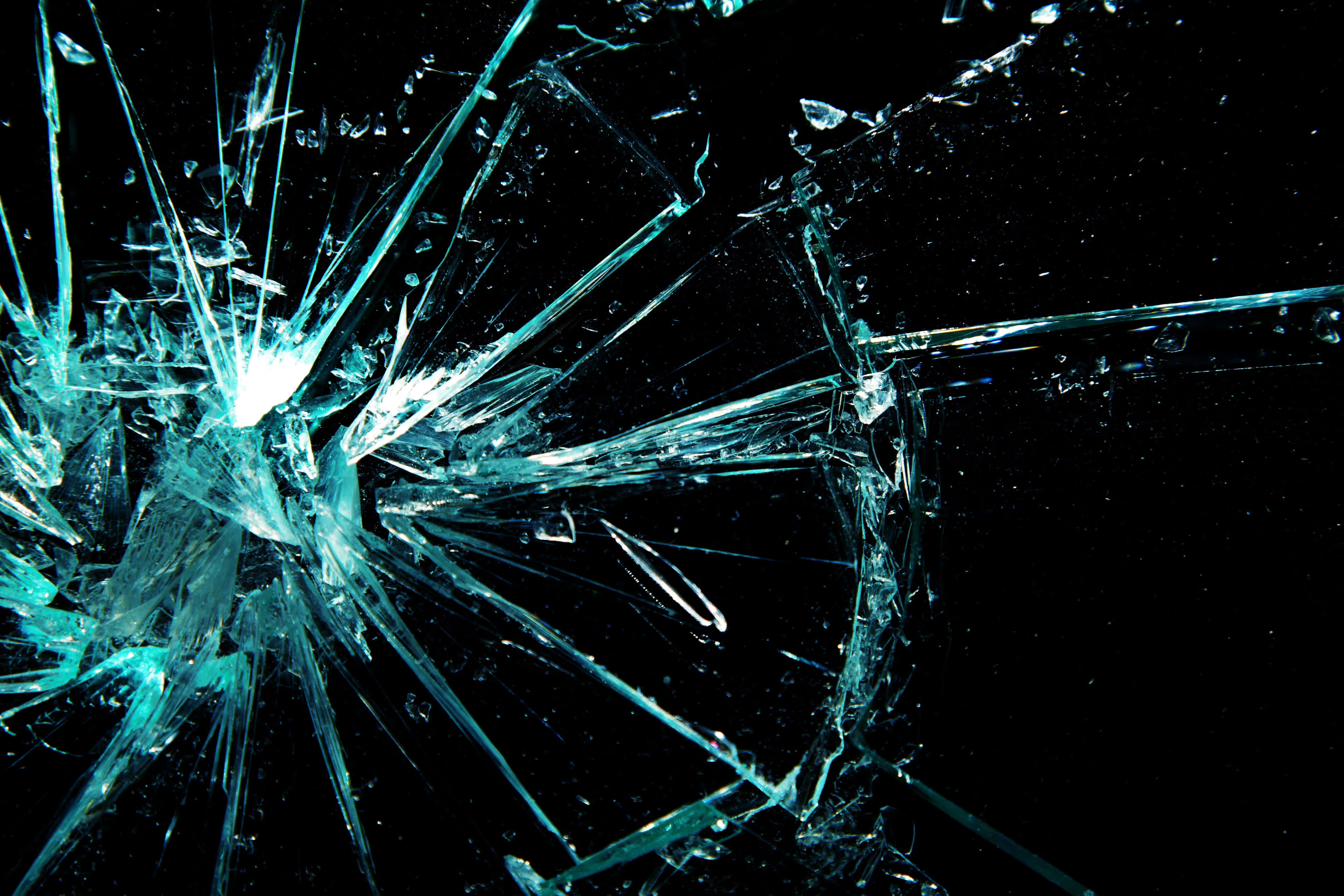 Broken Glass 桌布 壁紙 — 一塊破碎的玻璃表面，在黑色背景上，從明亮的中央撞擊點放射出尖銳的藍綠色裂紋。