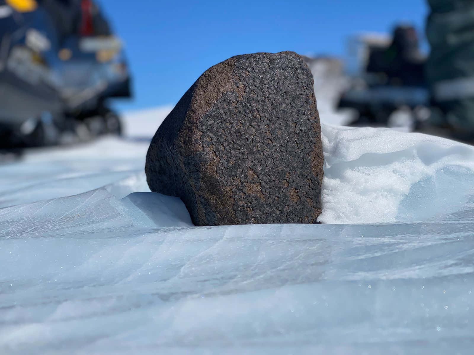 Antarctica 桌布 壁紙 — 一塊粗糙的深色岩石從紋理藍冰中突出，在晴朗的天空下，背景有模糊的雪地摩托車。