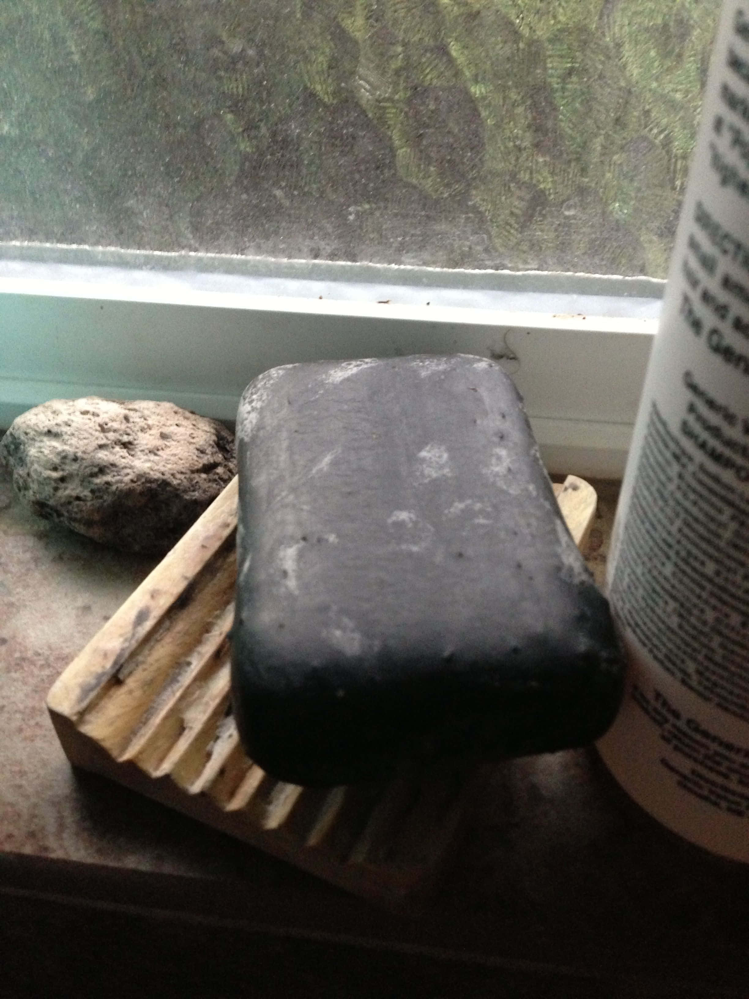Black Soap 桌布 壁紙 — 一塊經風化的黑色肥皂放在木製肥皂碟上，旁邊有一塊石頭和洗髮水瓶，窗台上可看到模糊的綠色景色。
