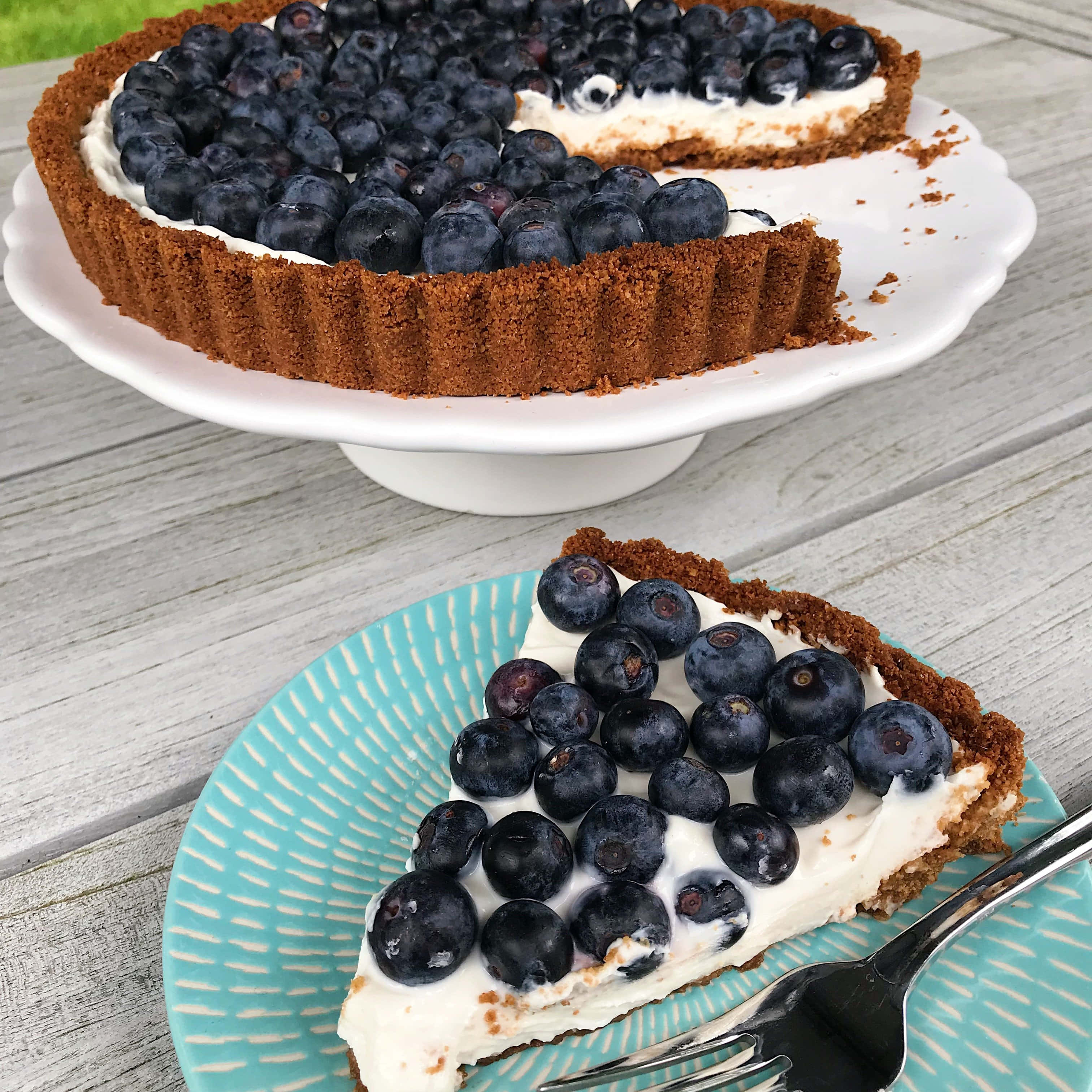 Blueberries Tart 桌布 壁紙 — 一塊藍莓撻放在青綠色碟子上，旁邊有一把叉，其餘的撻則放在白色蛋糕架上，置於一張風化的木桌上。