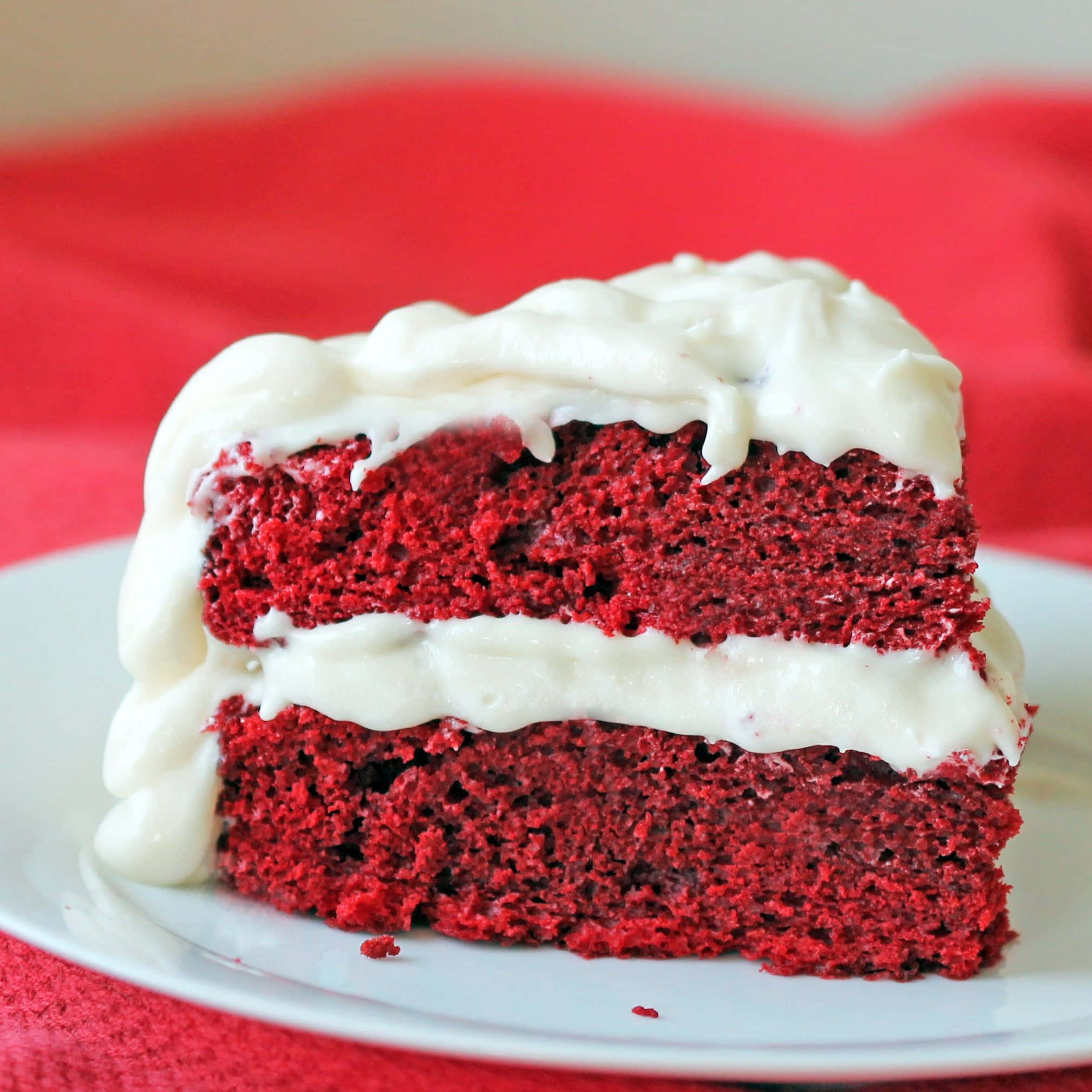 Red Velvet Cake 桌布 壁紙 — 一塊豐富的紅絲絨蛋糕片，帶有奶油白色糖霜，在白色碟子上滴落邊緣，背景為紅色布料。