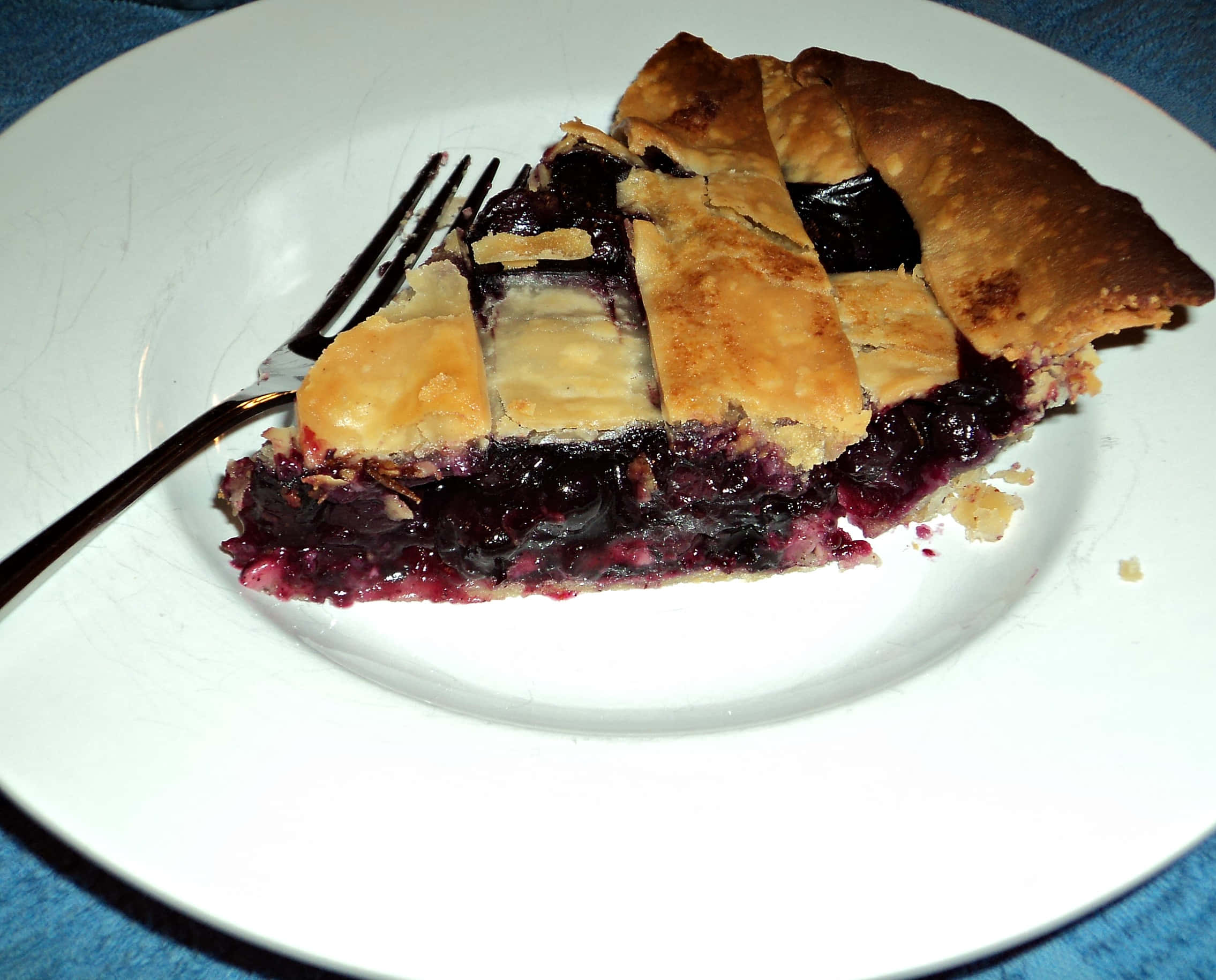 Blueberry Pie 桌布 壁紙 — 一塊金黃色格子頂藍莓批，內餡多汁，放在白色碟上，附有叉子。