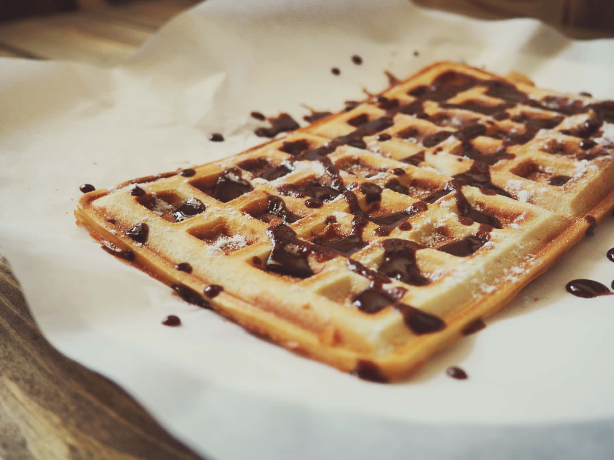 Waffle 桌布 壁紙 — 一塊金黃色的窩夫淋上濃郁的朱古力醬並撒上糖粉，放在牛油紙上。