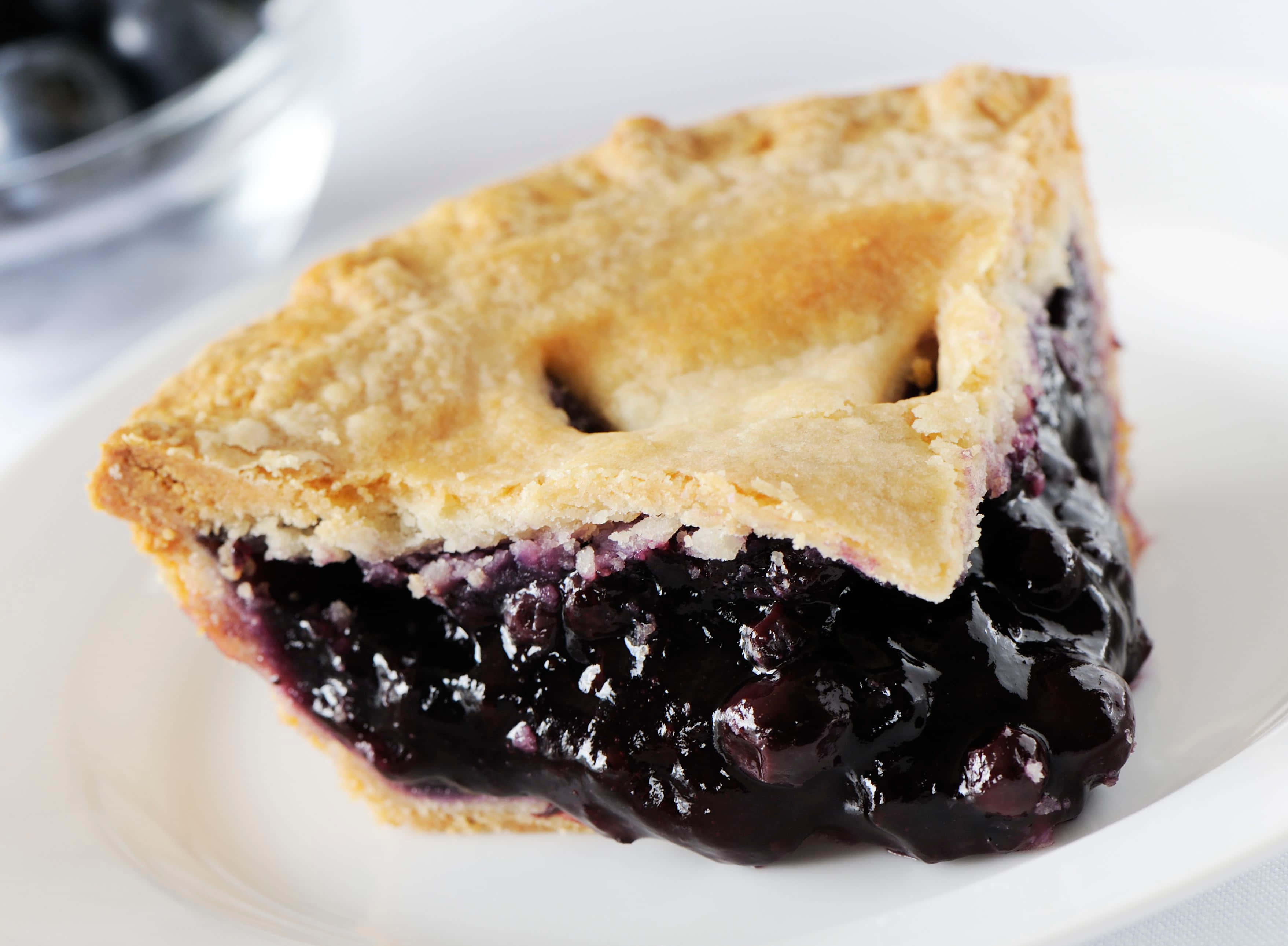 Blueberry Pie 桌布 壁紙 — 一塊金黃酥脆的藍莓批，帶有 juicy 深色餡料，放在白色碟上，背景有一碗新鮮莓果。