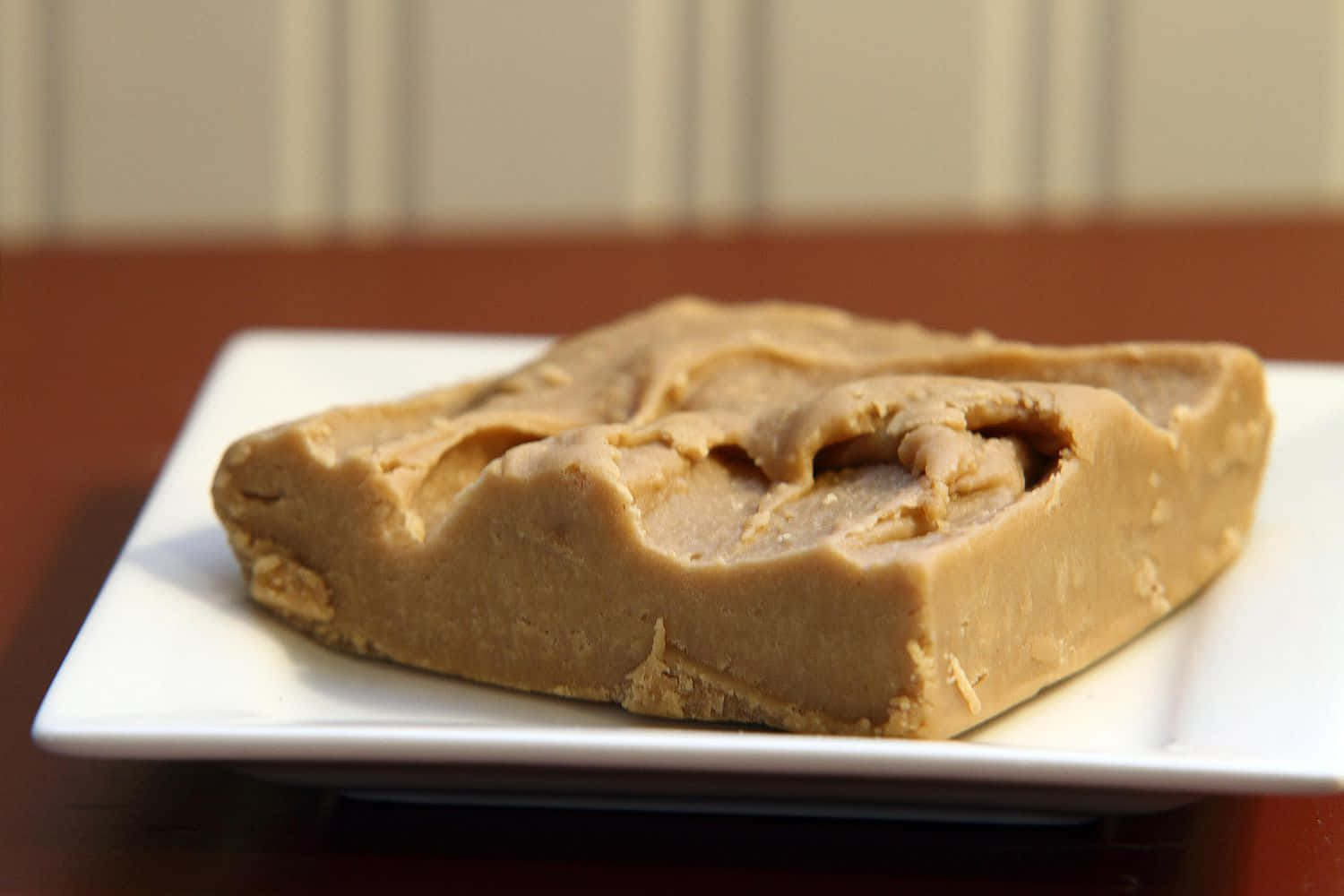 Penuche Fudge 桌布 壁紙 — 一塊順滑的花生醬軟糖片放在木桌上的白色碟子上，背景是米色鑲板牆。